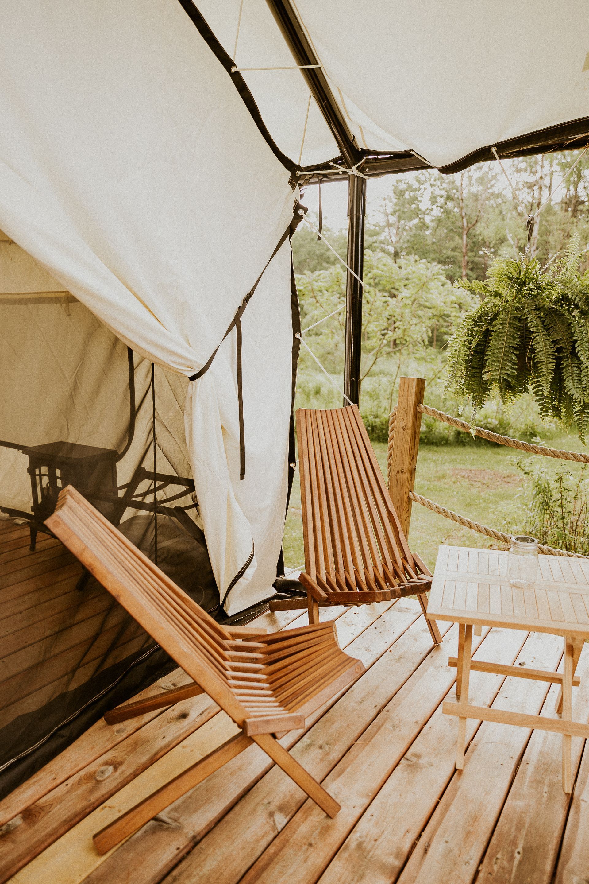 Wooden chairs on a porch of a tent-like structure, with a lush green backdrop.
