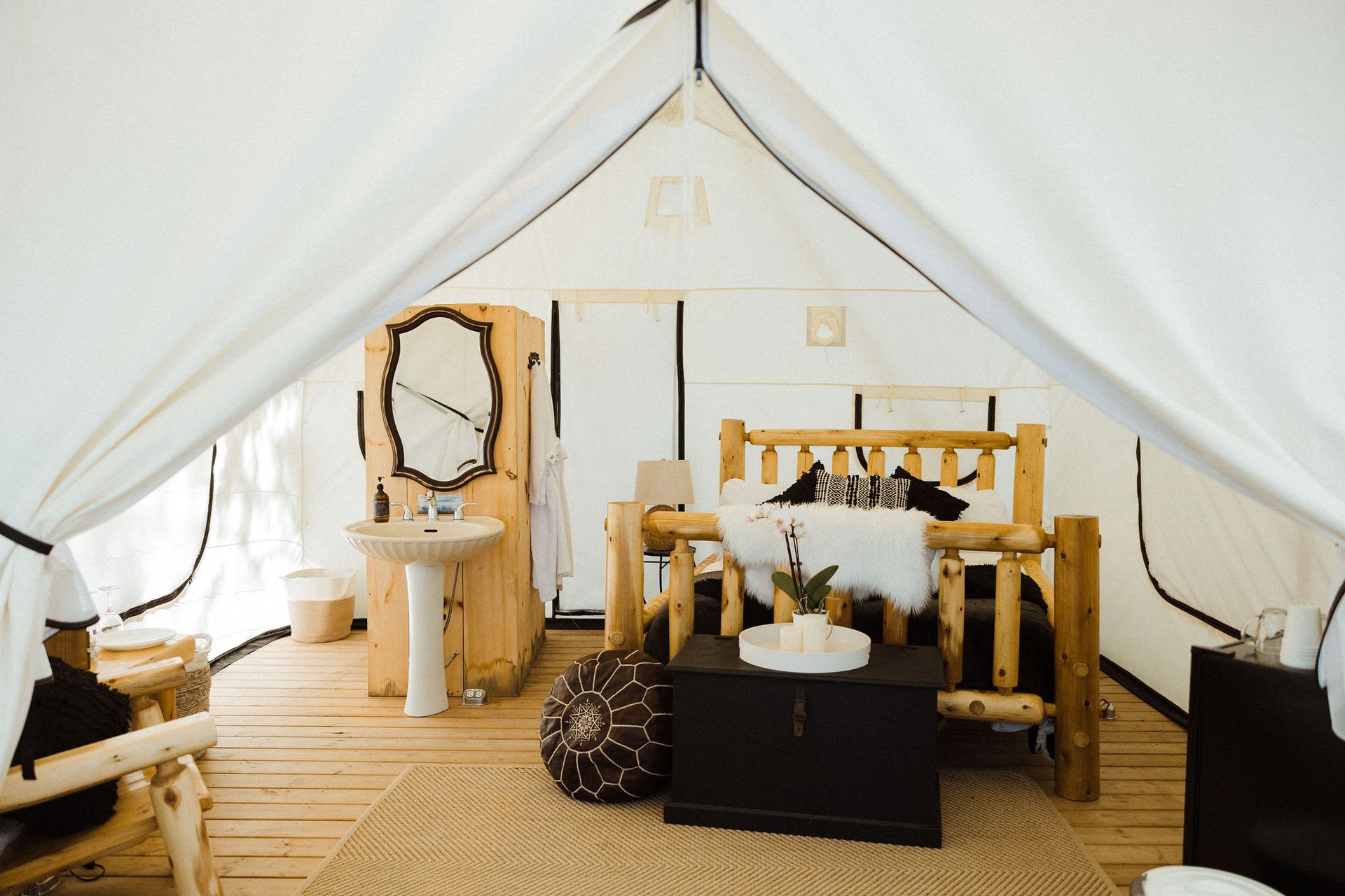 Interior of a glamping tent with bed, sink, mirror, and wooden furniture on a wood floor.