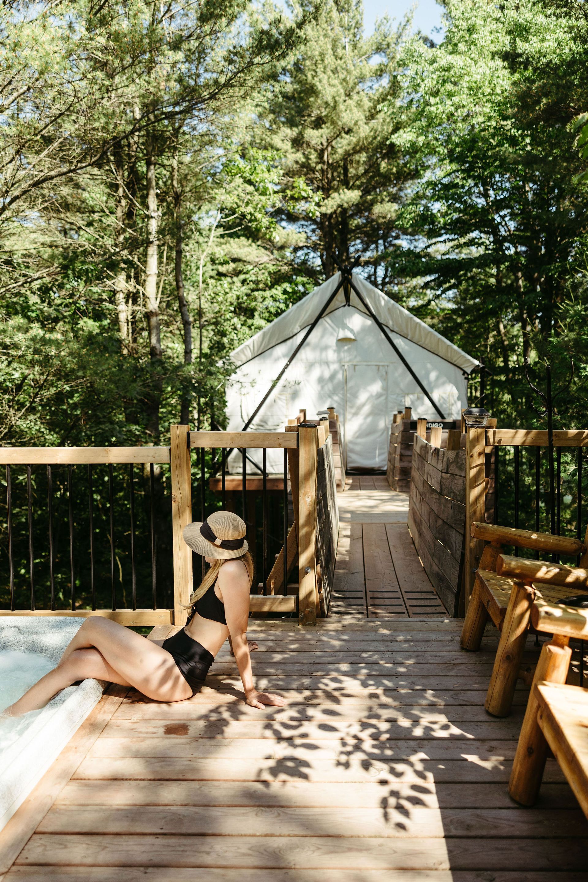 Woman in swimsuit relaxes by a hot tub on a wooden deck with a tent in the background, surrounded by trees.