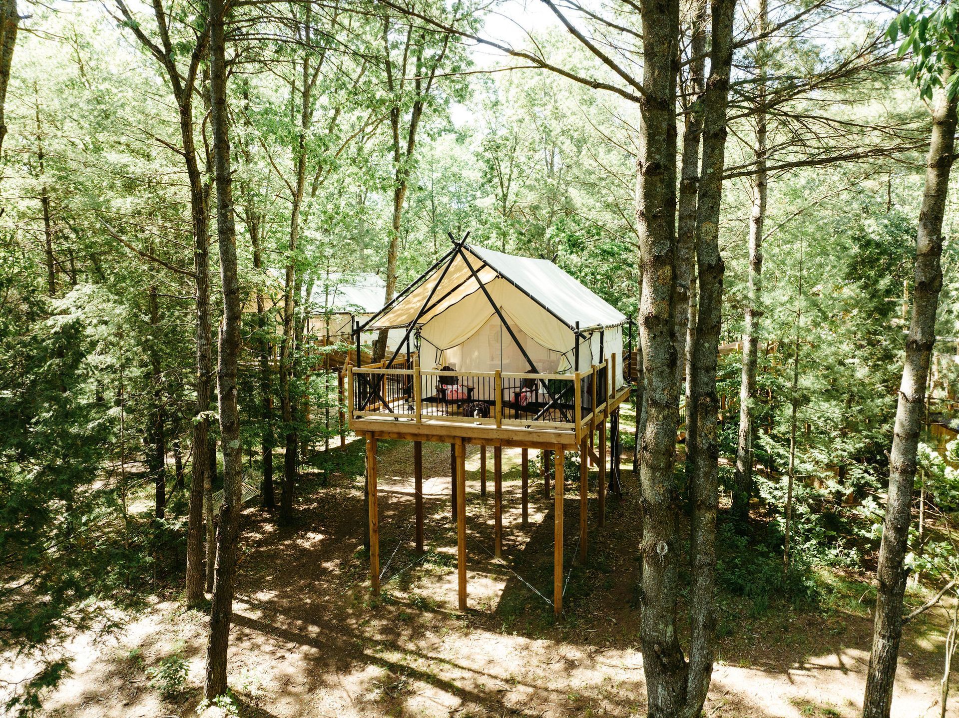 A canvas-walled cabin on stilts in a forest. It has a deck with chairs.