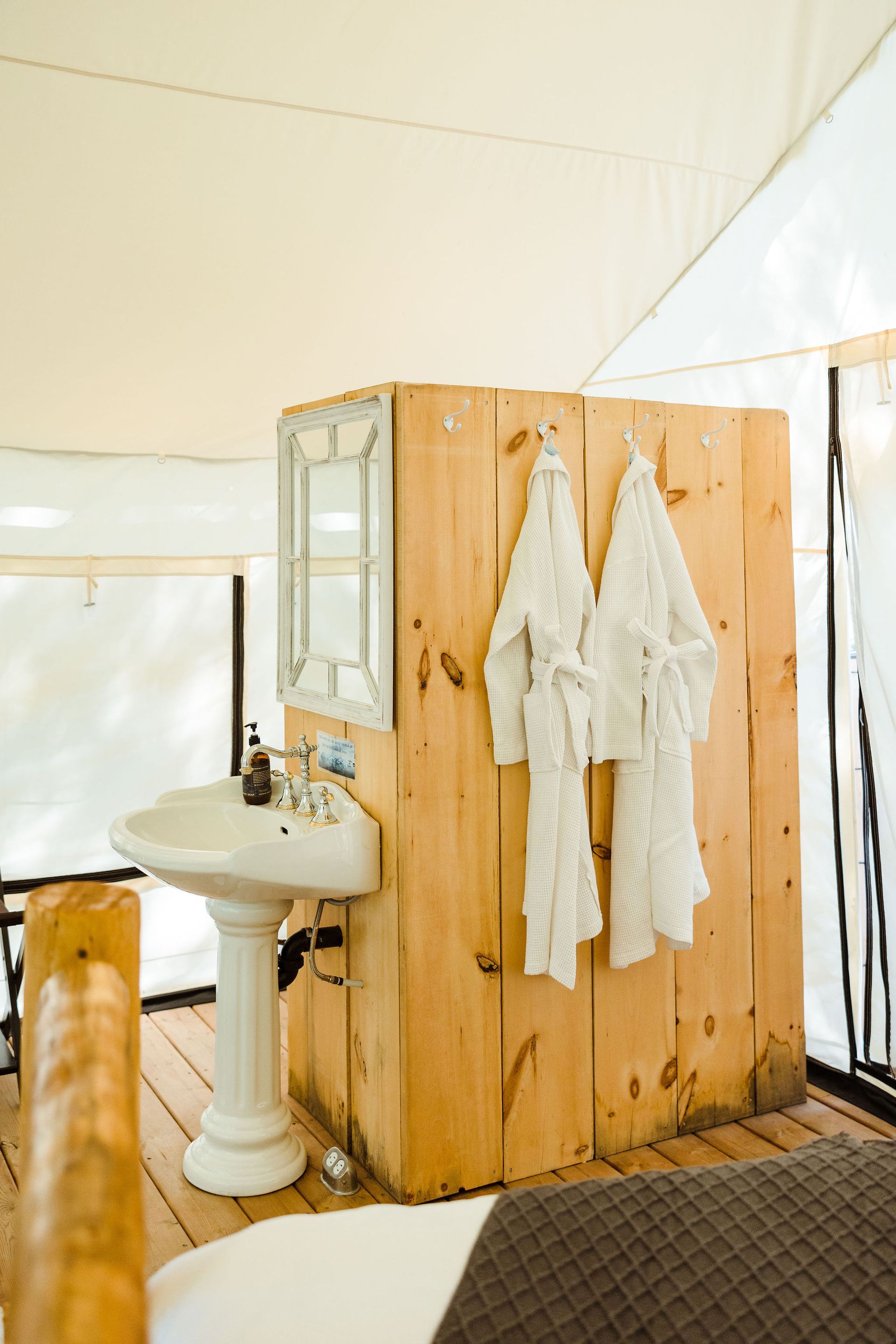 Rustic bathroom in a tent: pedestal sink, wooden walls, robes hanging, mirror, light tent ceiling.