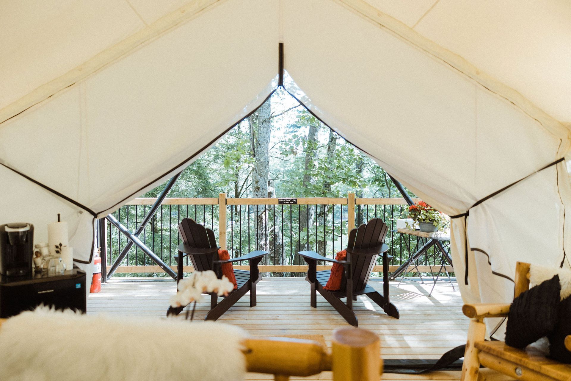 Interior of a glamping tent with wooden chairs on a deck overlooking trees. Coffee maker and decor visible.