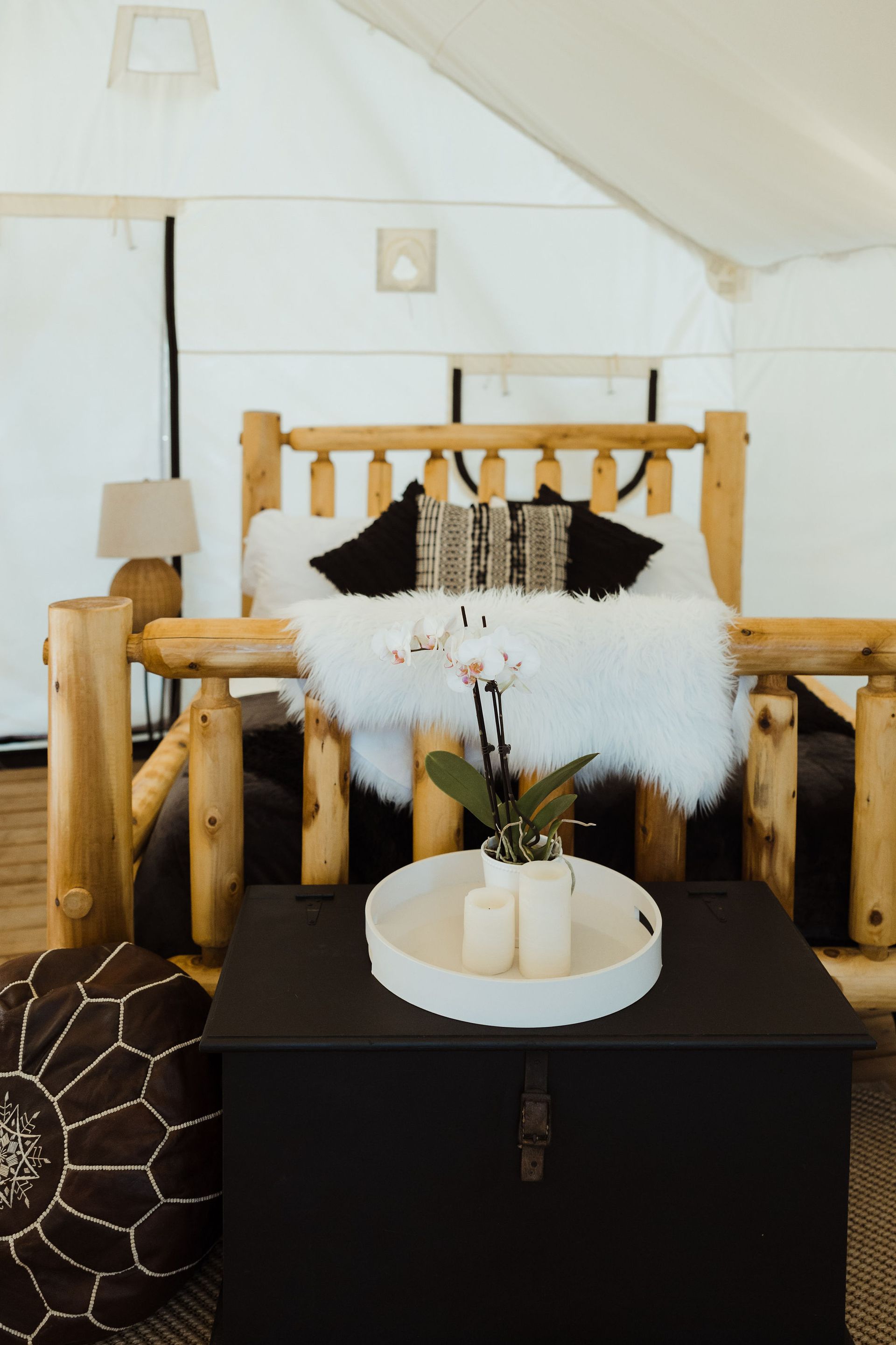 Glamping tent interior: Wooden bed with white fur, black storage chest, flower, and decorative pillows.