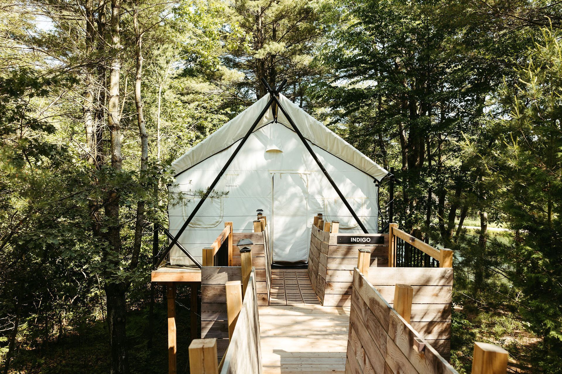 White tent structure in a forest, accessed by a wooden ramp and stairs.