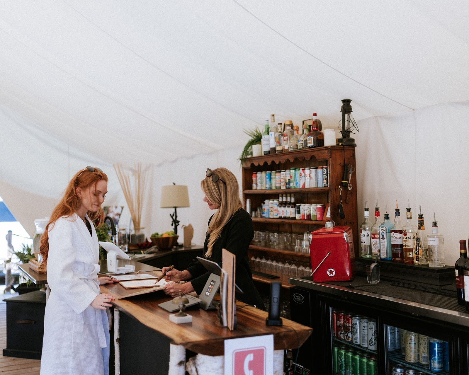 Two women at a bar with a wooden counter and shelves of drinks, inside a white tent.