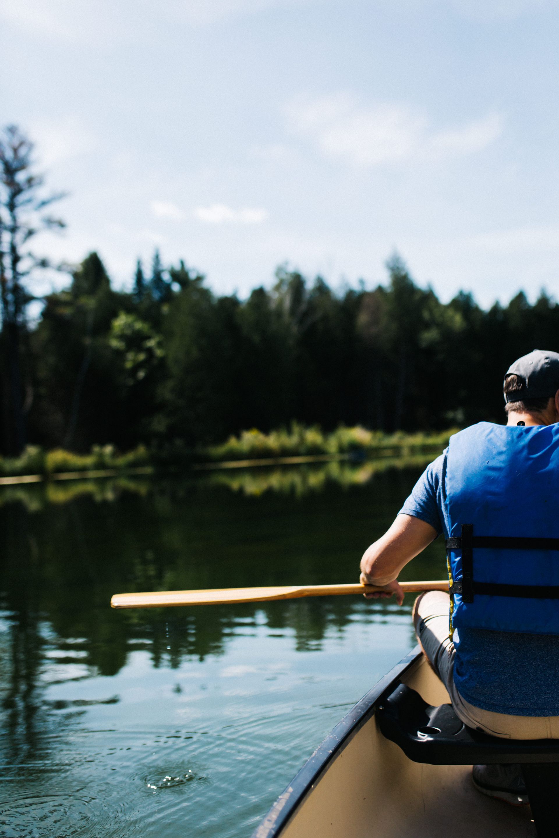 Person paddles a canoe on a lake, wearing a blue life vest. Trees line the far shore on a sunny day.