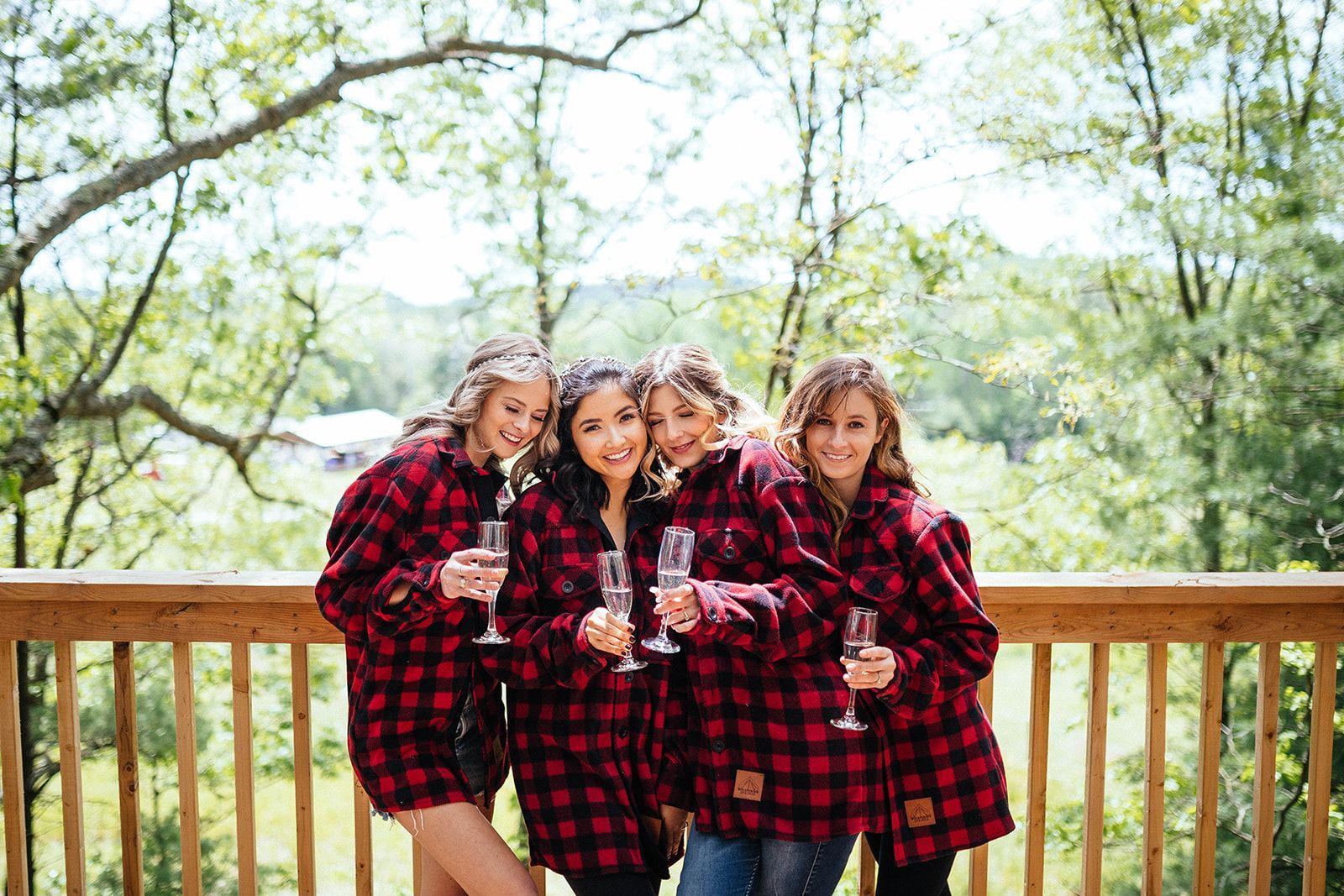 Four women in red and black plaid shirts, holding champagne flutes on a wooden deck.