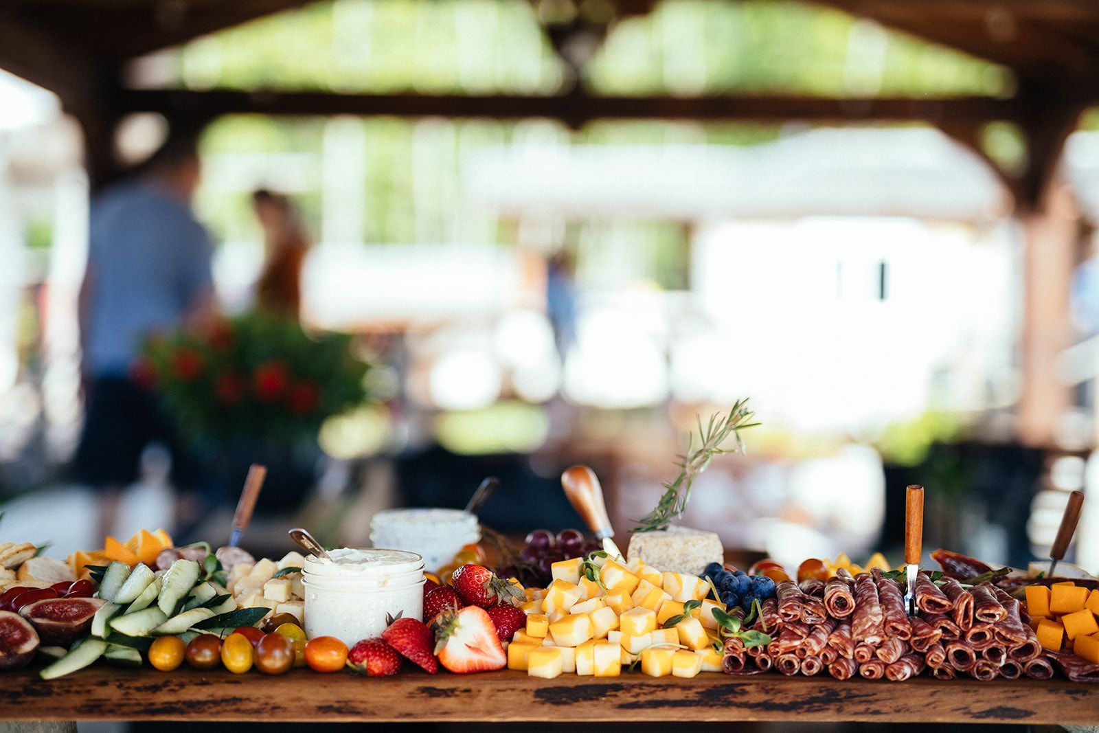 Charcuterie board: cheeses, fruits, and meats arranged on a table outdoors; people blurred in background.