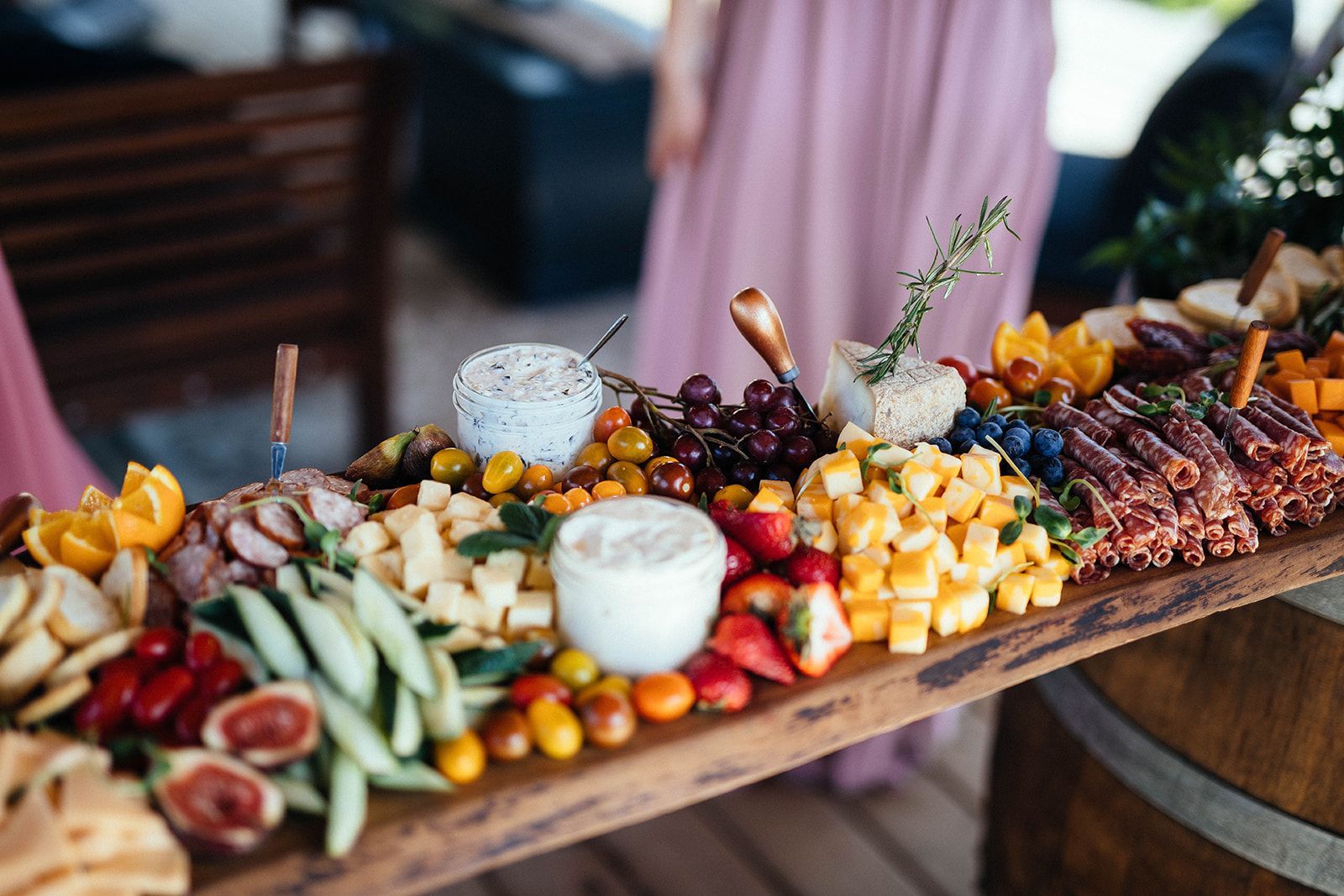 Charcuterie board on a wooden table, featuring cheese, fruit, and meat, with a person in a pink dress in the background.