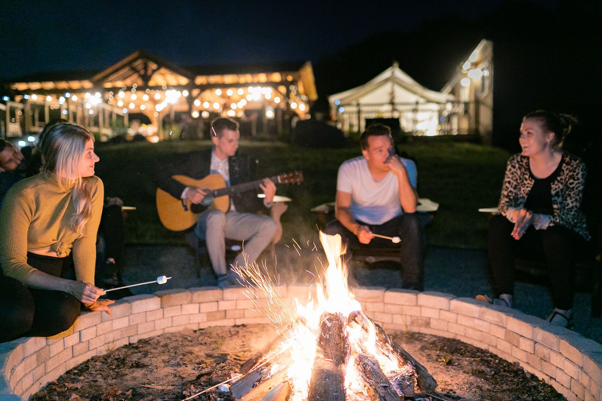 People around a bonfire, one playing guitar, at night. Building in background, marshmallow roasting.