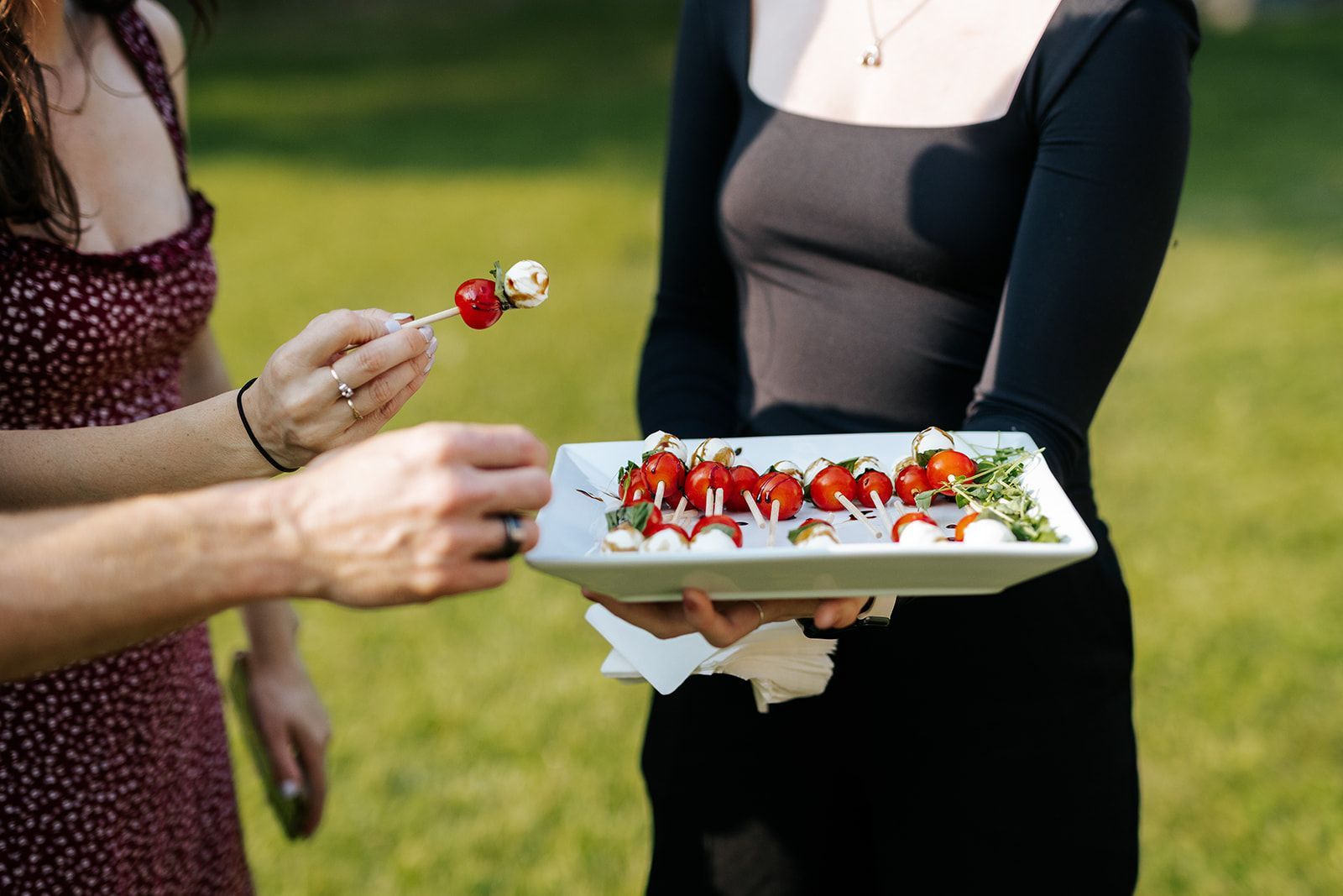 Woman in black dress holds a tray of tomato and mozzarella skewers; another person takes a skewer. Outdoors, sunny.