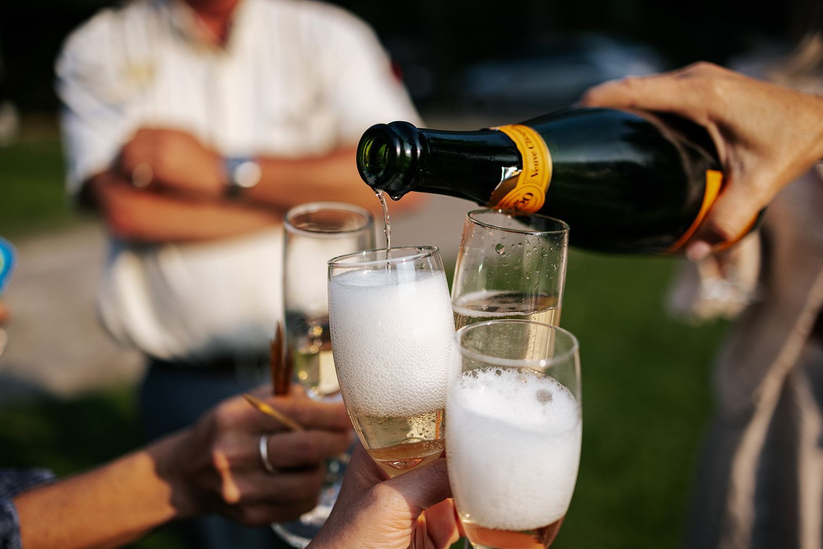 Champagne being poured into fluted glasses at an outdoor gathering; someone's arm reaches to hold the bottle.