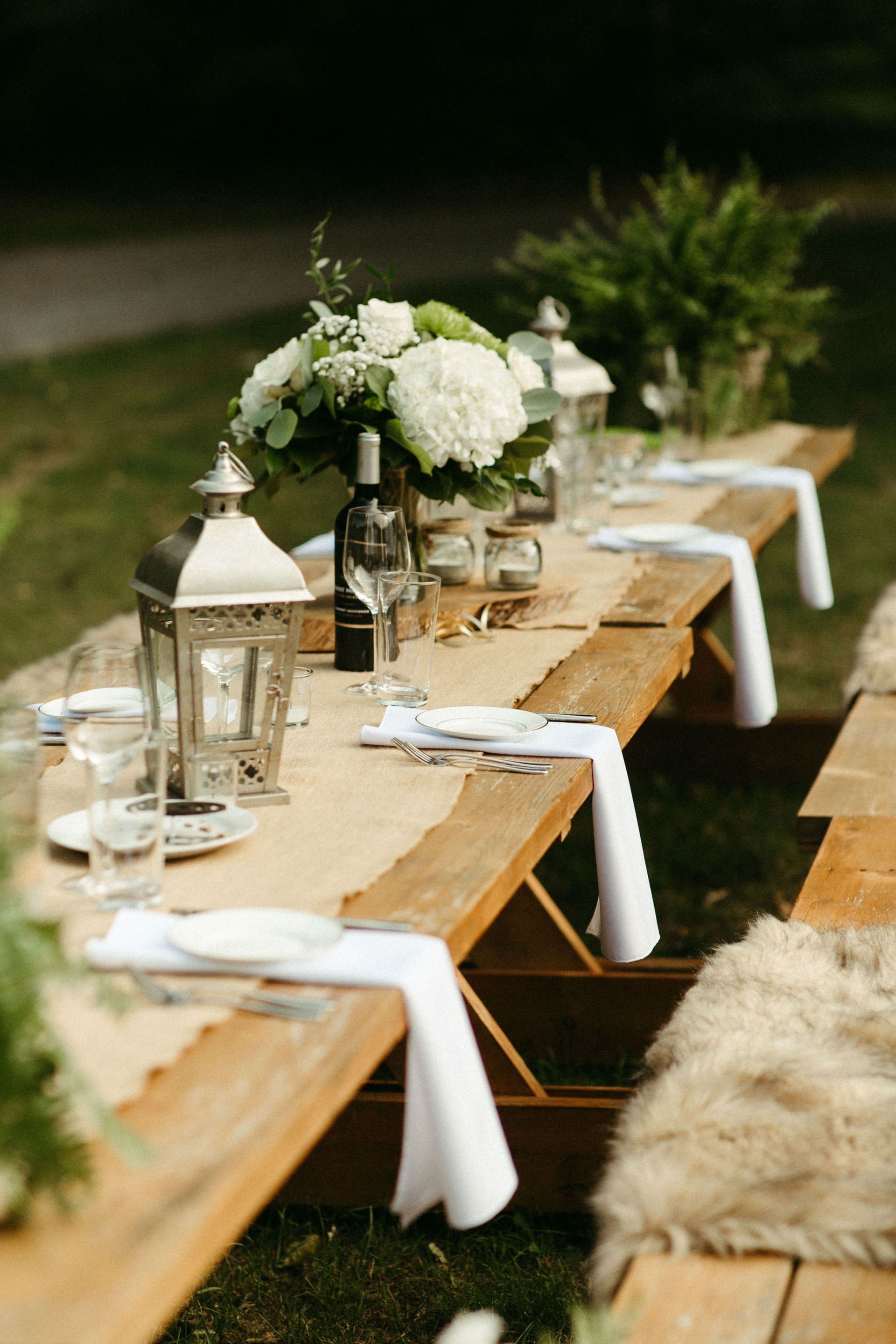 Outdoor dining table set with white flowers, lanterns, and wine. Wooden bench seating on grass.
