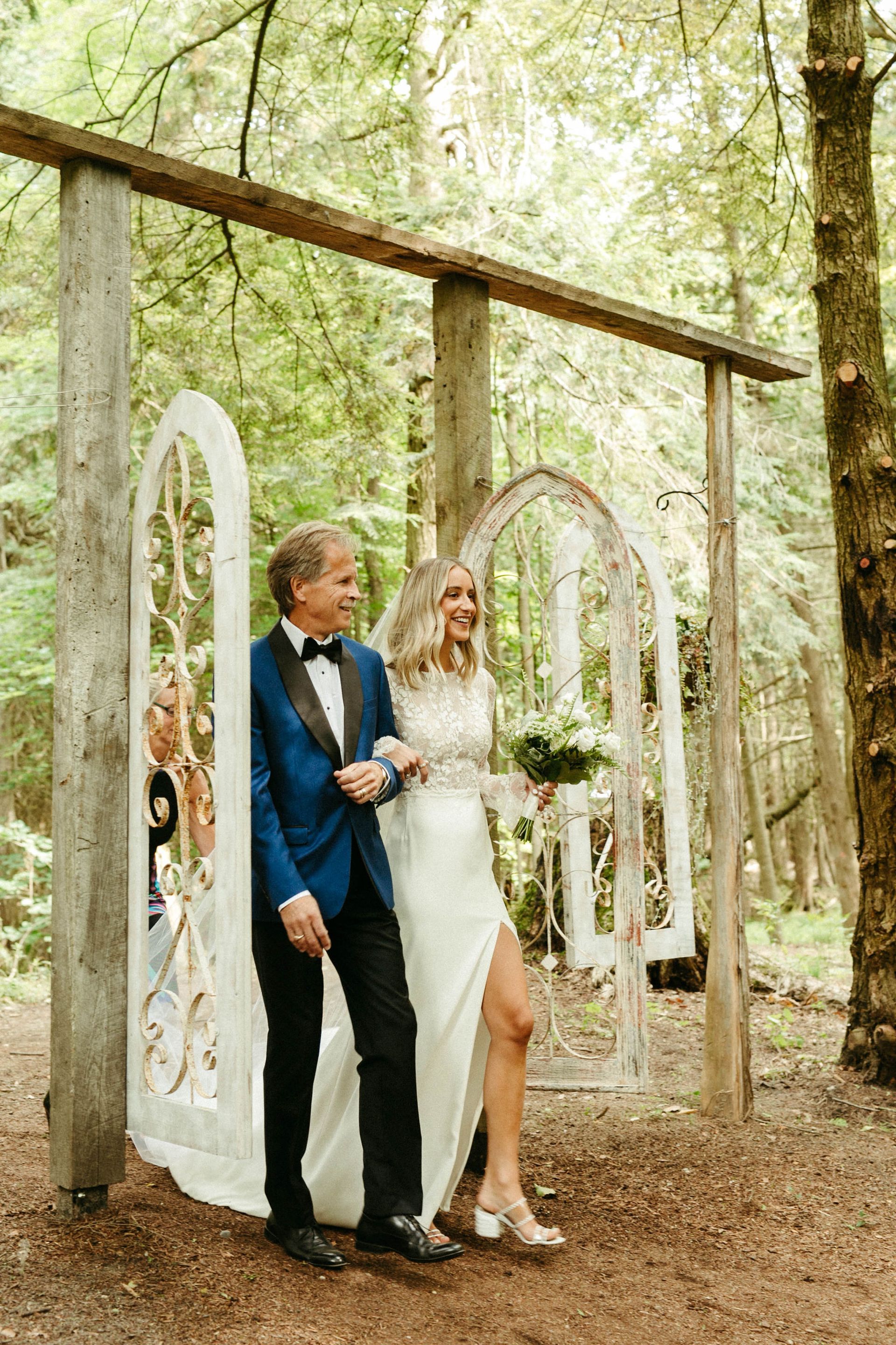 Bride in white dress walks down an aisle in the woods, arm in arm with a man in a blue suit.