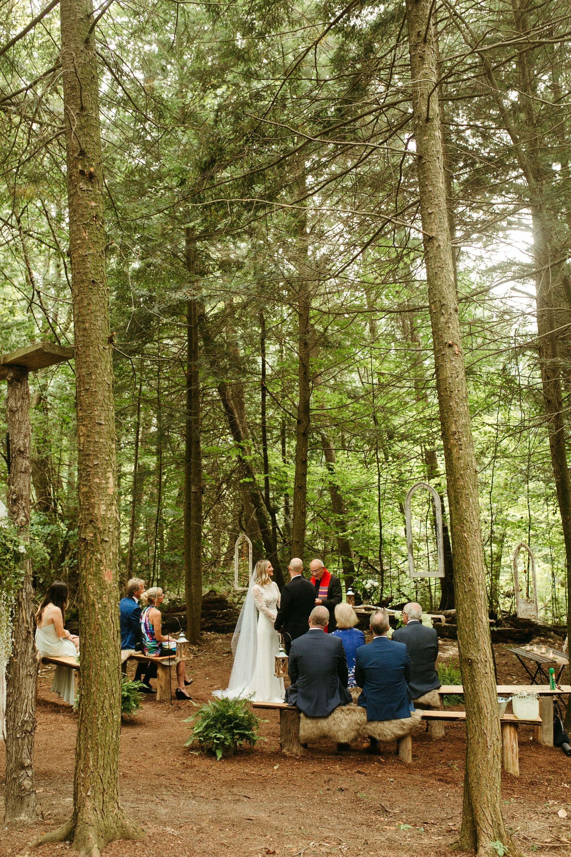 Wedding ceremony in a forest setting. Bride and officiant stand before seated guests.