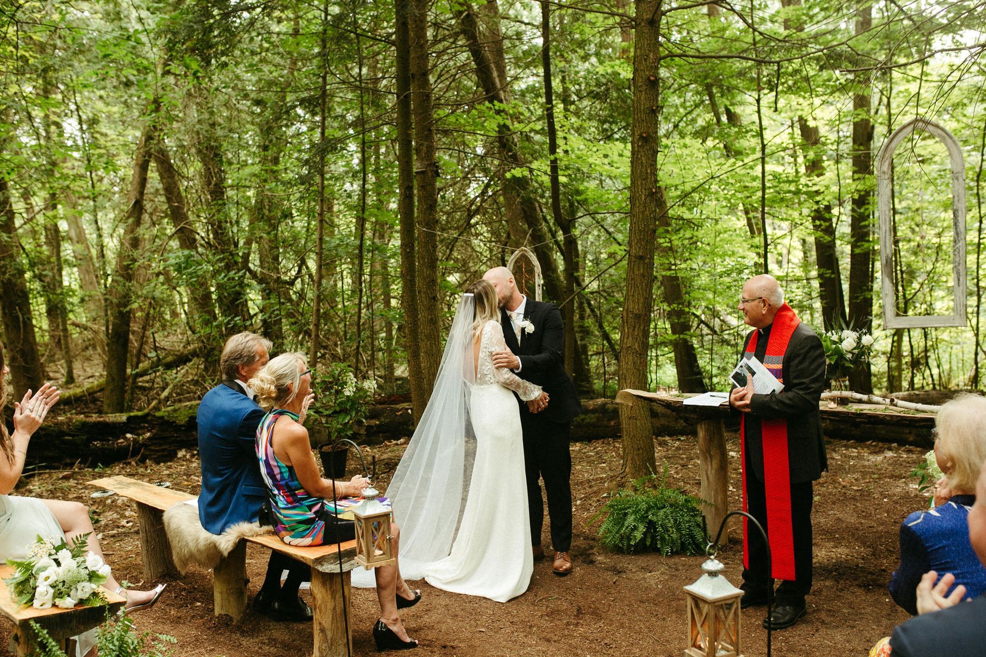 Wedding ceremony in a forest clearing: couple kissing, officiant, guests seated on benches.