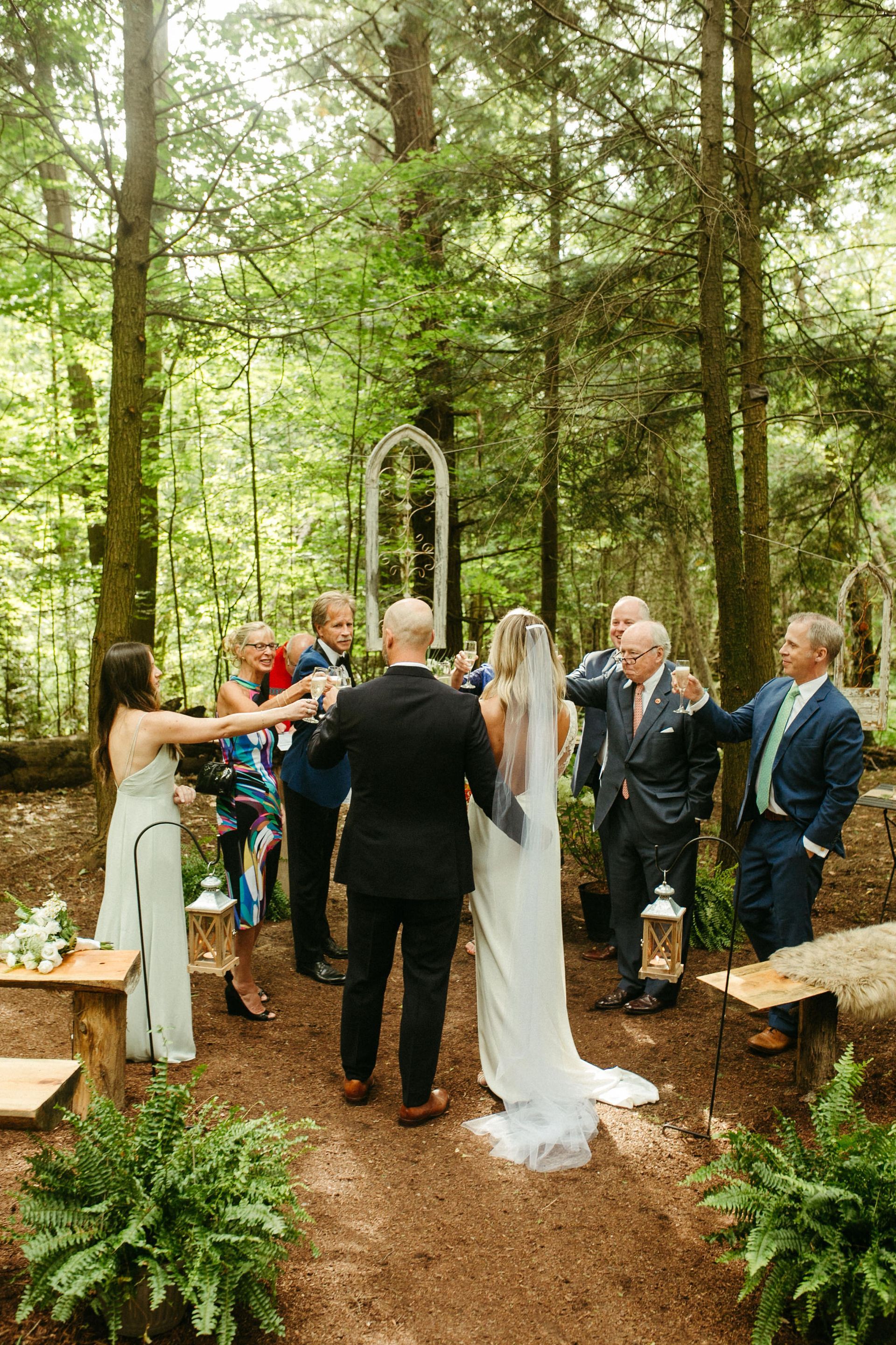 Wedding ceremony in a forest setting. Bride and groom with guests, toasting. Trees and a decorative archway.