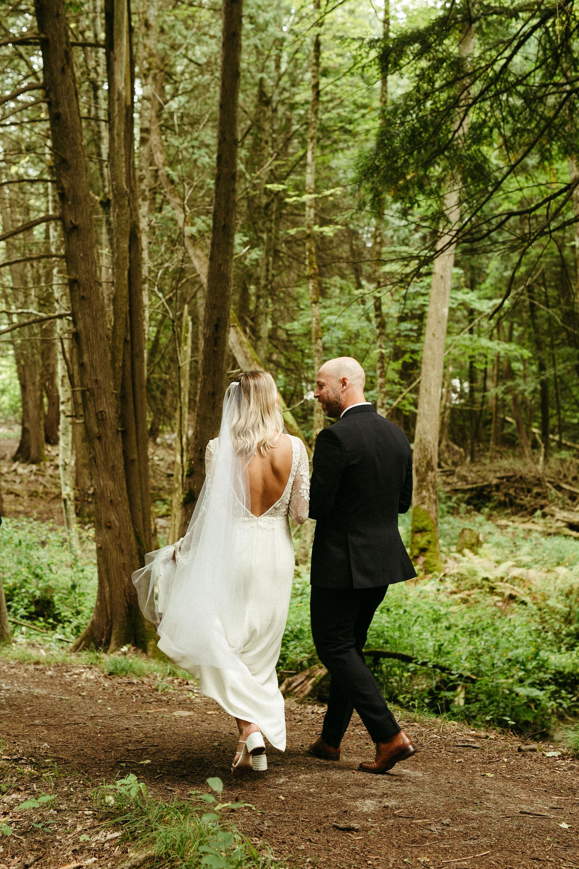 Bride and groom walk through a forest on a dirt path, wedding attire.