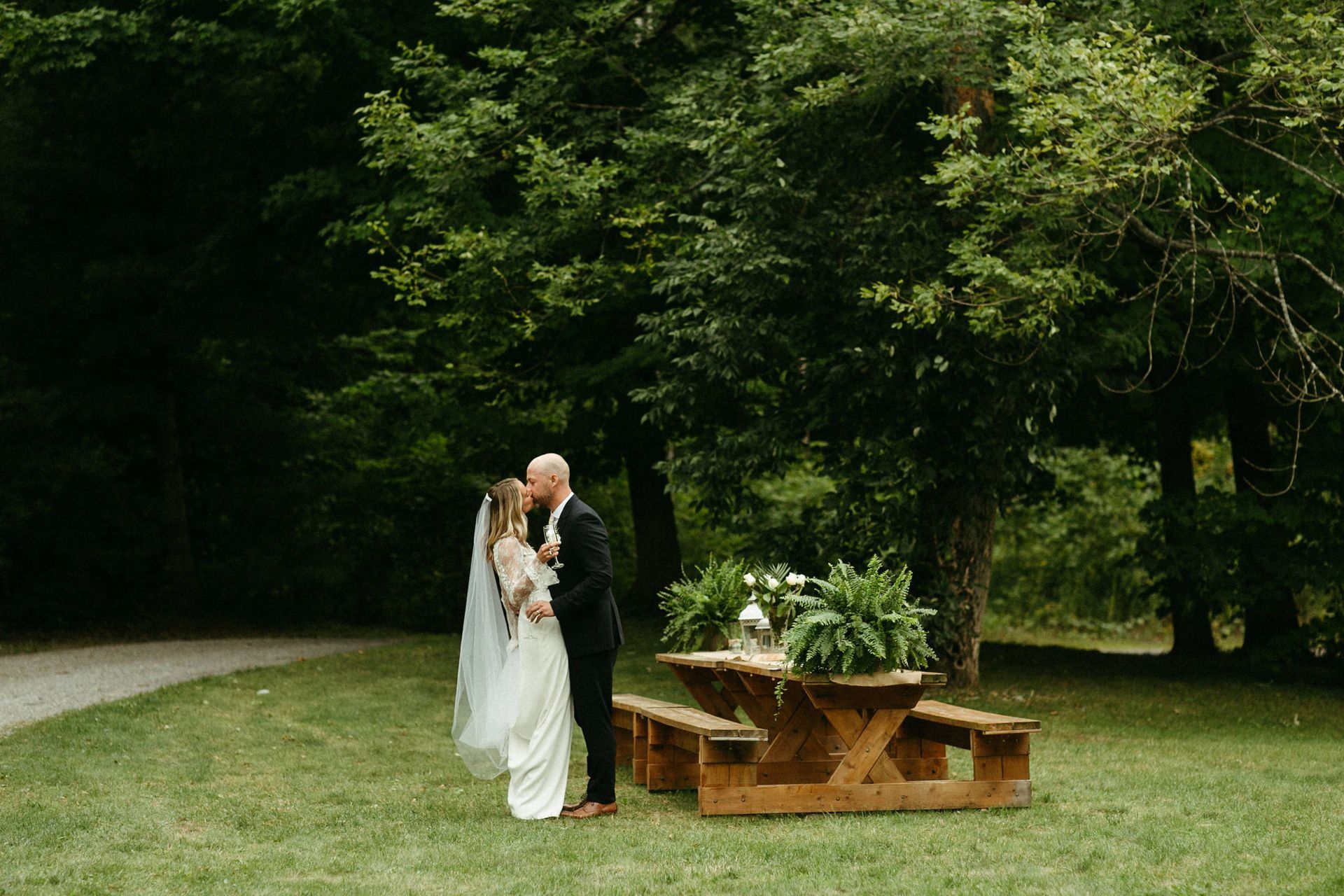Couple kissing at outdoor wedding ceremony near a picnic table with greenery.