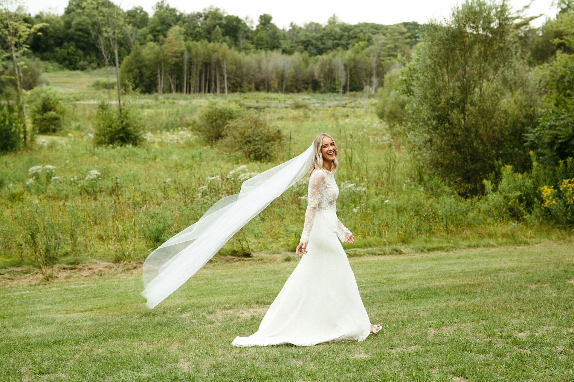Bride in white gown and long veil smiles, walking in a grassy field with trees in the background.