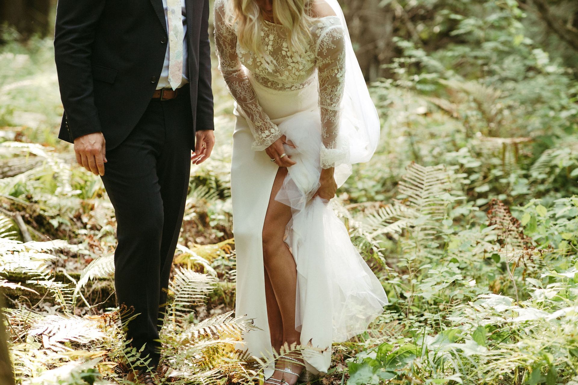 Bride lifting wedding dress, revealing garter, with groom in forest.