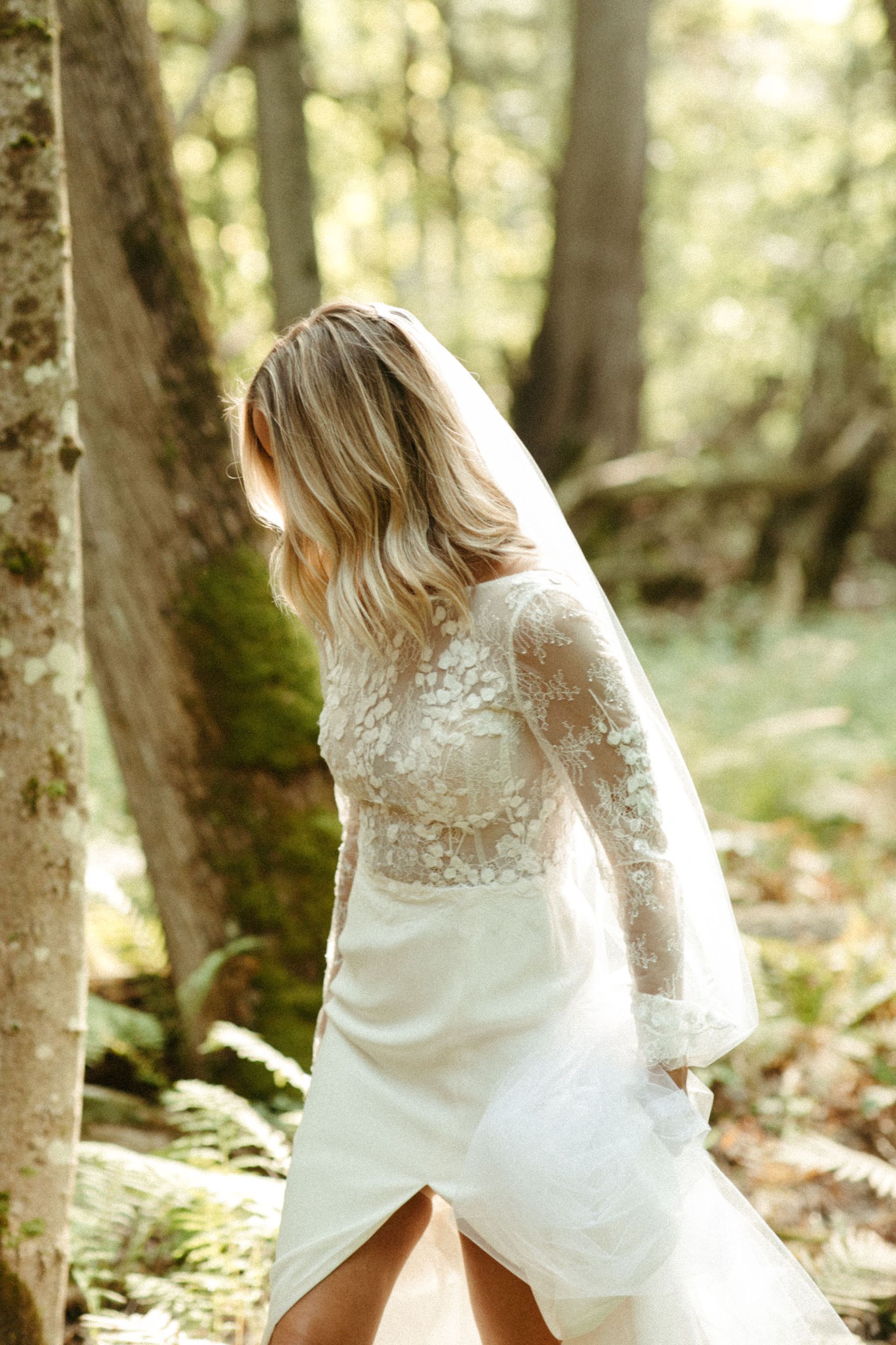 Bride in lace wedding dress walks through forest, holding the skirt, with veil and trees.