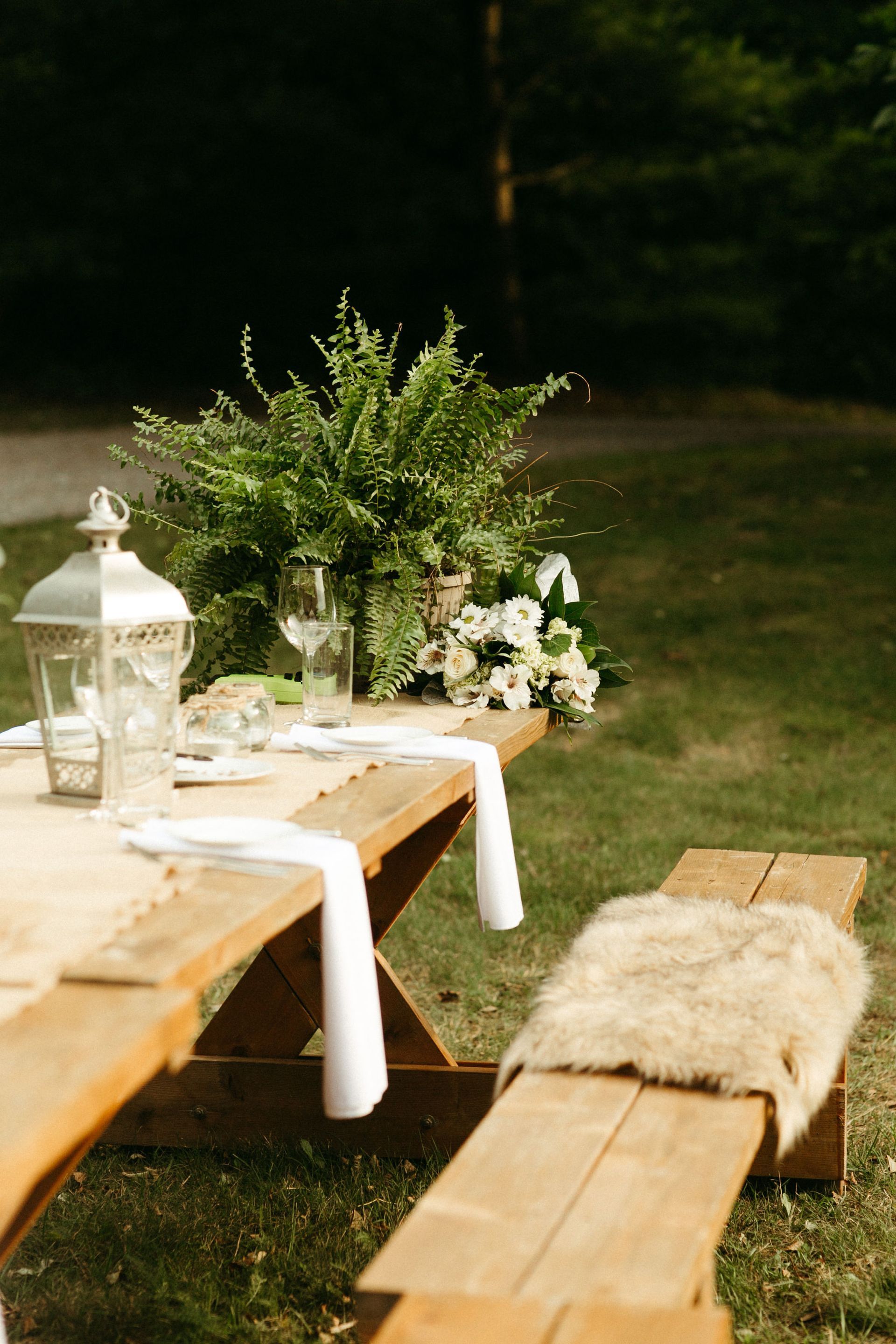 Picnic table set for an outdoor event with greenery, white linens, and a fur bench seat.