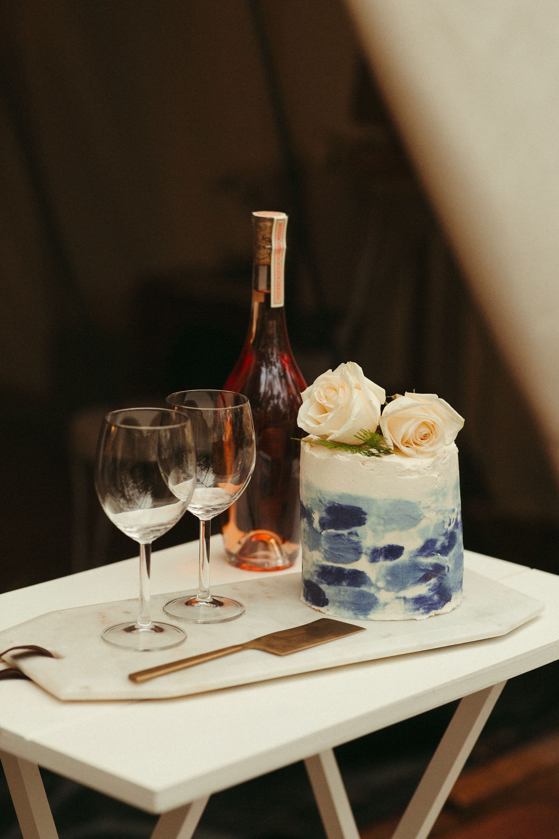 Cake, wine, and glasses on a white table. Blue watercolor cake with roses. Wine bottle in background.