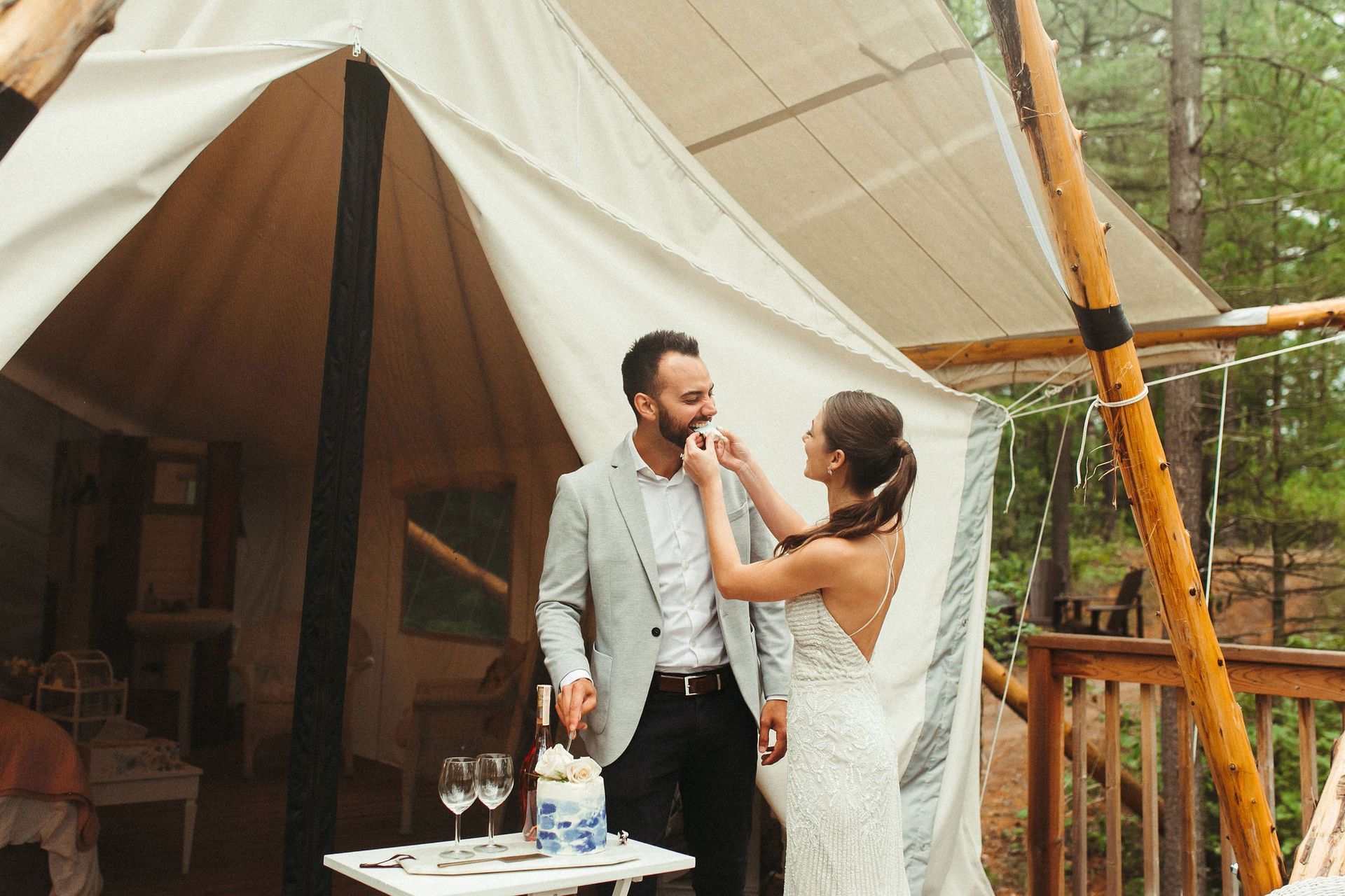 Bride feeding cake to groom on a wooden deck near a glamping tent.