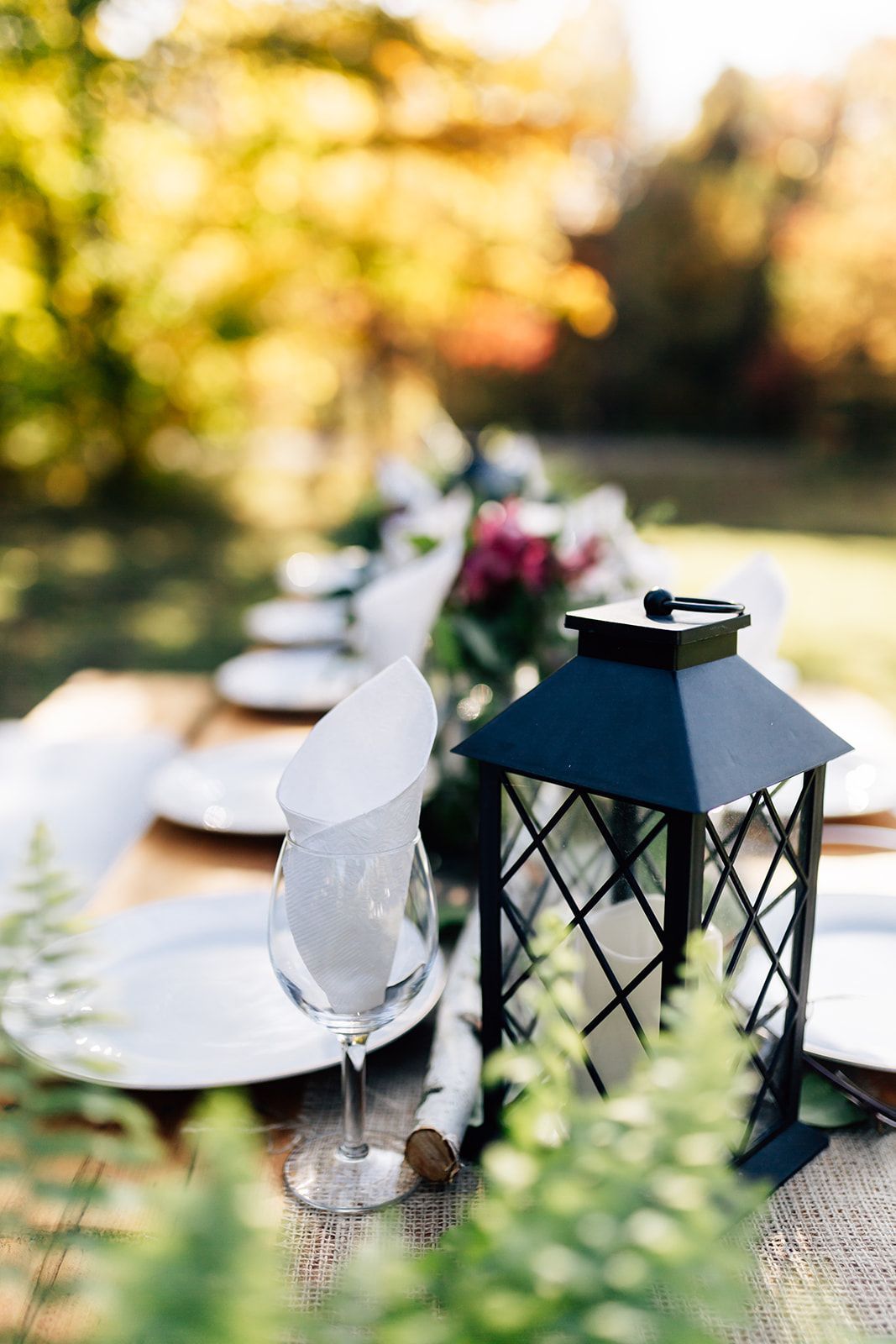 Outdoor table setting with lantern, wine glass, and floral arrangement against a blurred fall background.