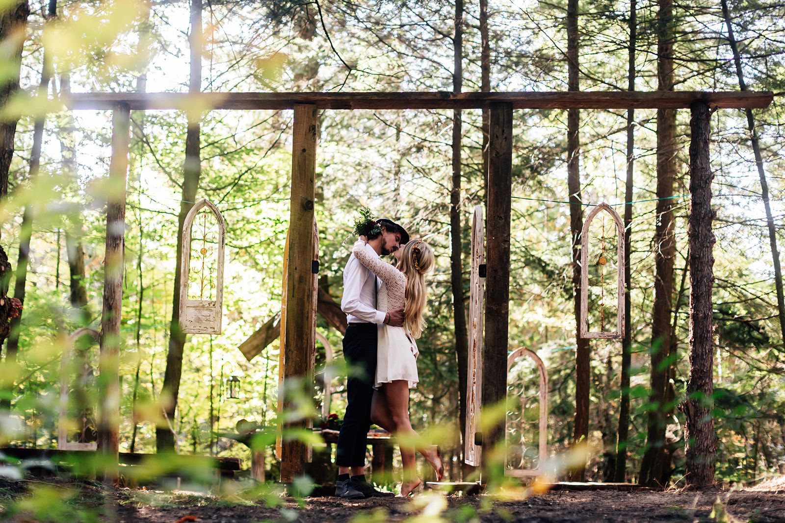 Couple kissing in a forest, standing beneath a wooden arch. Sunny, with greenery and trees surrounding them.