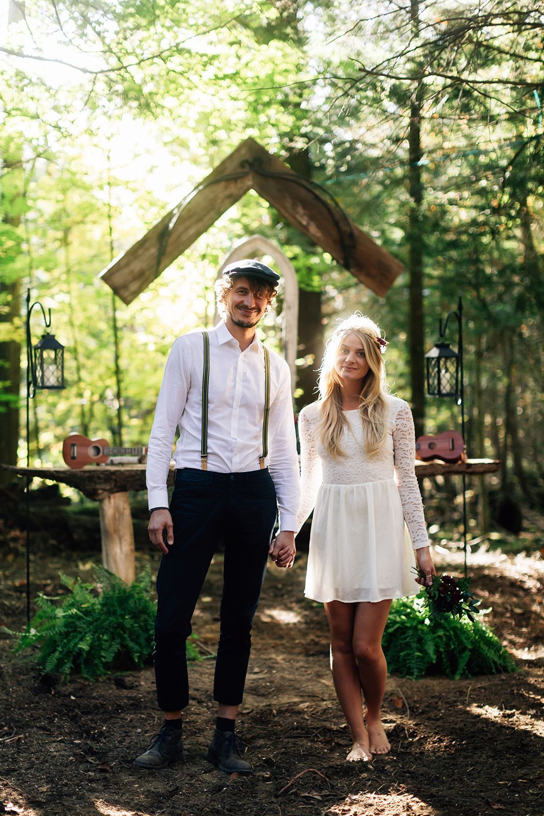 Couple holding hands in a forest, standing near a wooden archway; the woman is barefoot, the man wearing suspenders.