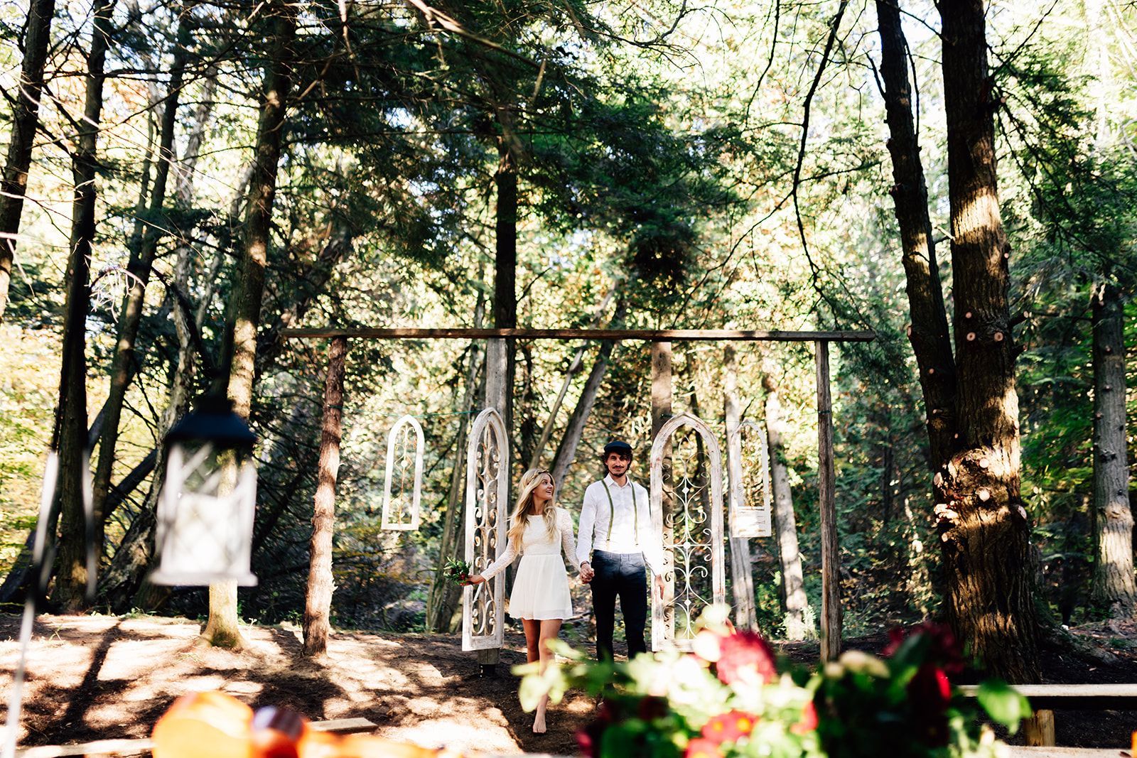 Couple standing under a wooden archway decorated with white arches in a forest clearing; ceremony setting.
