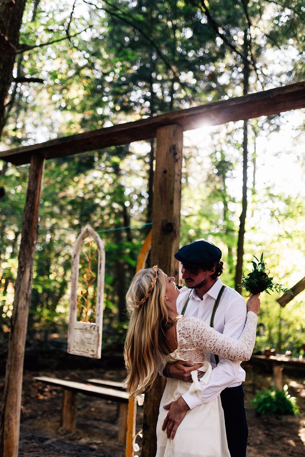 Couple embraces outdoors; woman in white dress, man in hat; sunlight shines through trees.
