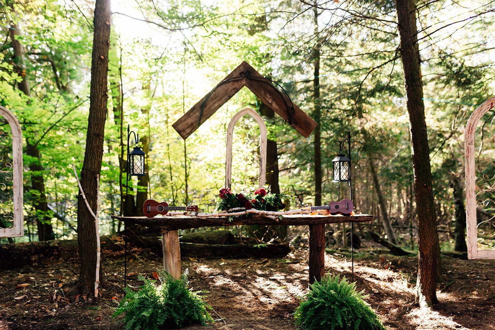 Wedding altar in a forest setting: wooden table with floral arrangement, decorative arches, sunlight through trees.