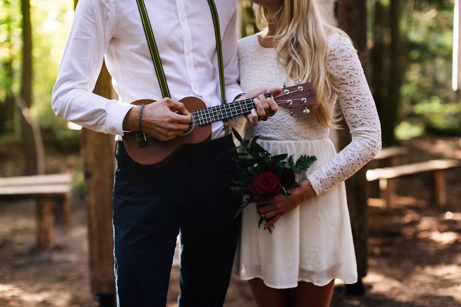 Man in suspenders plays ukulele, woman holds flowers, embracing in woods.