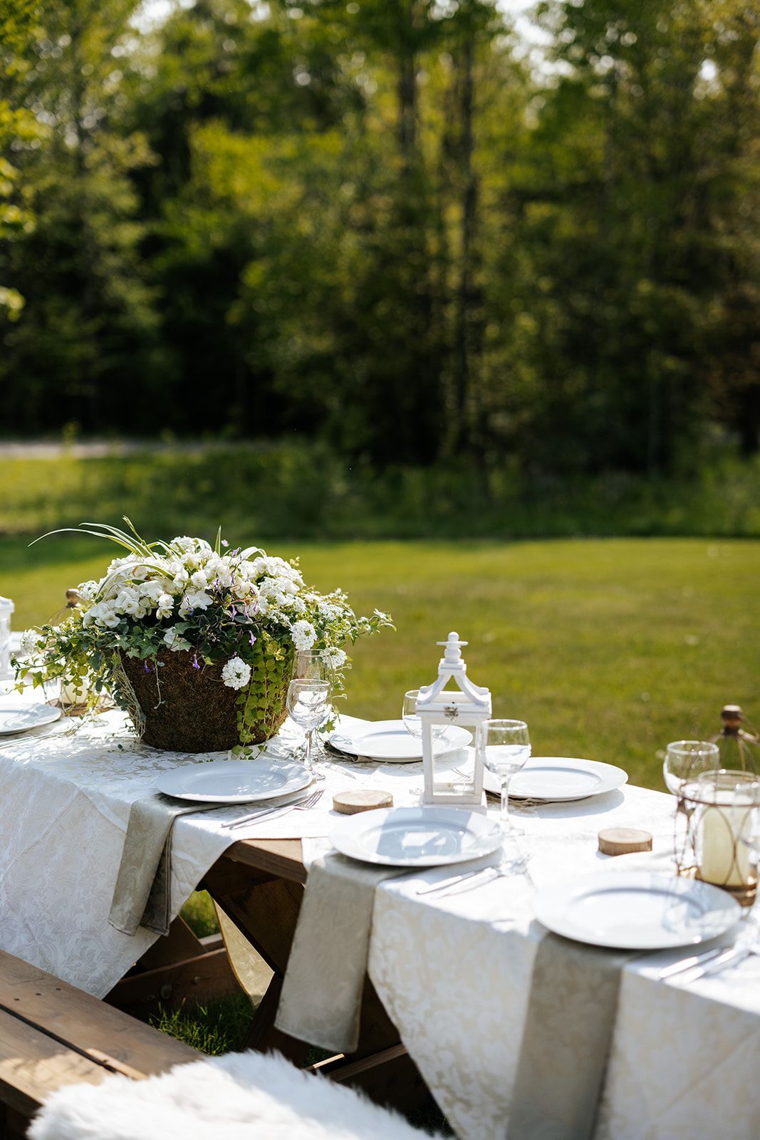 Outdoor dining table set with white dishes, floral centerpiece, and lanterns against a green lawn.