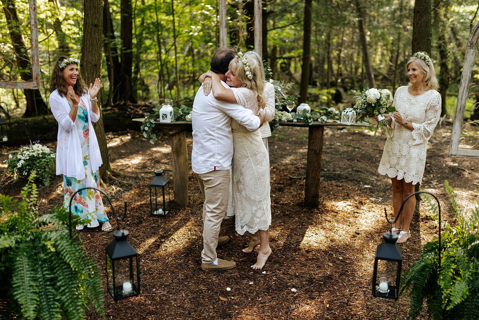 Couple embracing after wedding ceremony in forest, officiant clapping, bridesmaid smiling, altar.