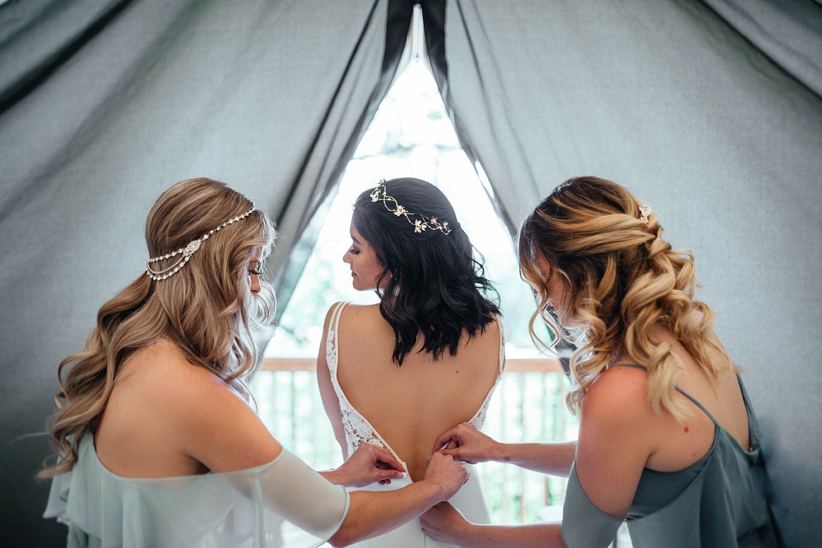 Bridesmaids helping a bride fasten her wedding dress outdoors.