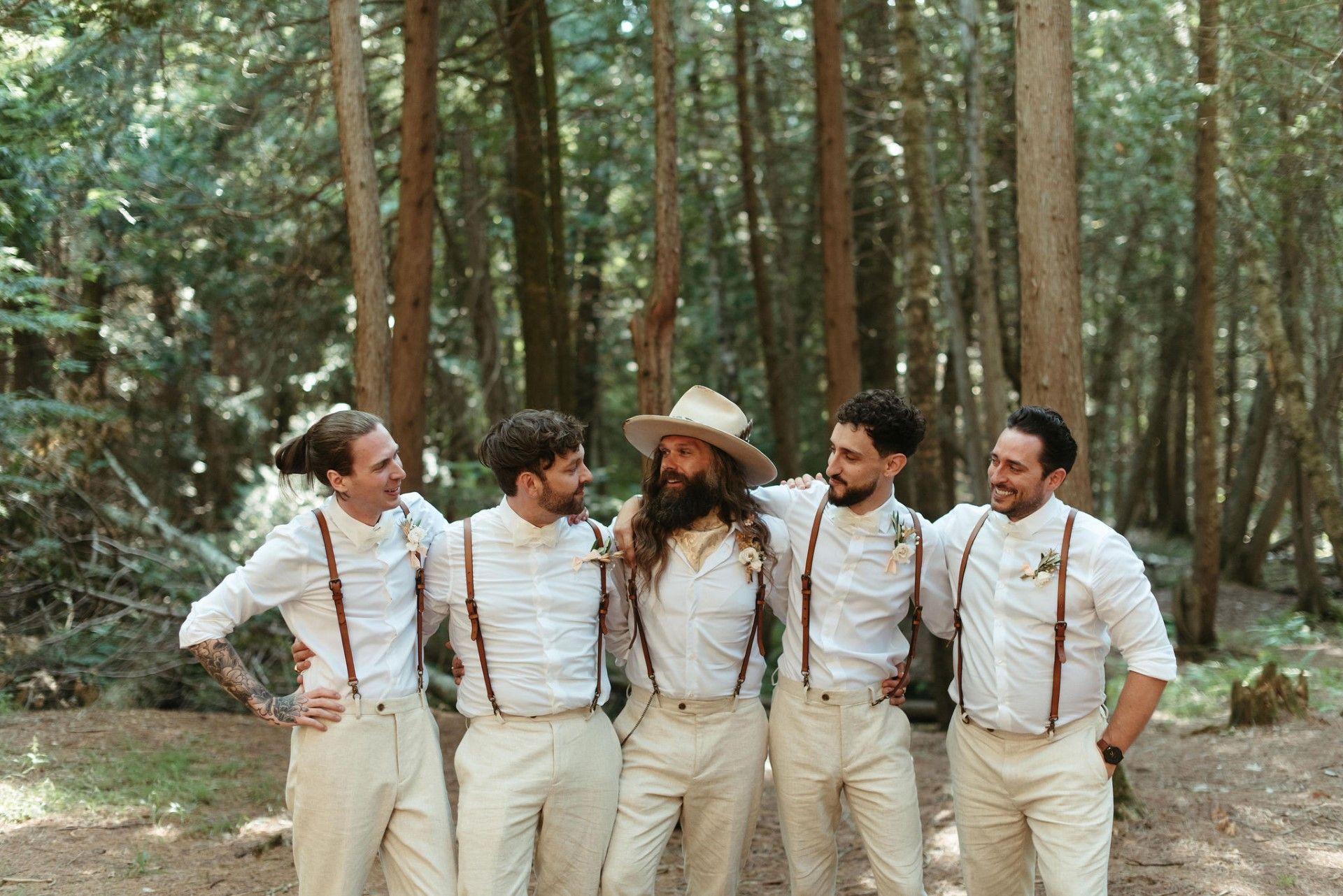 Five men in tan pants, white shirts, and suspenders pose in a forest. One wears a hat, and they smile.