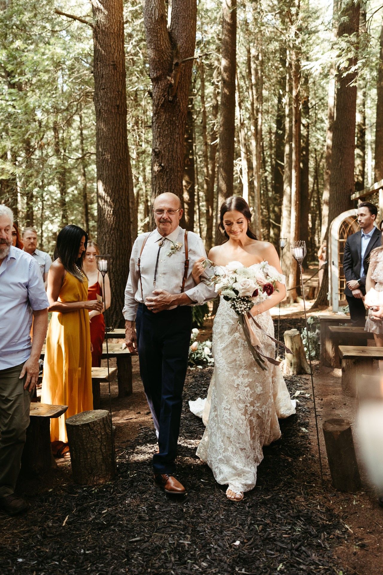 Bride walks down forest aisle with father, holding flowers. She wears a white gown. Attendees watch.