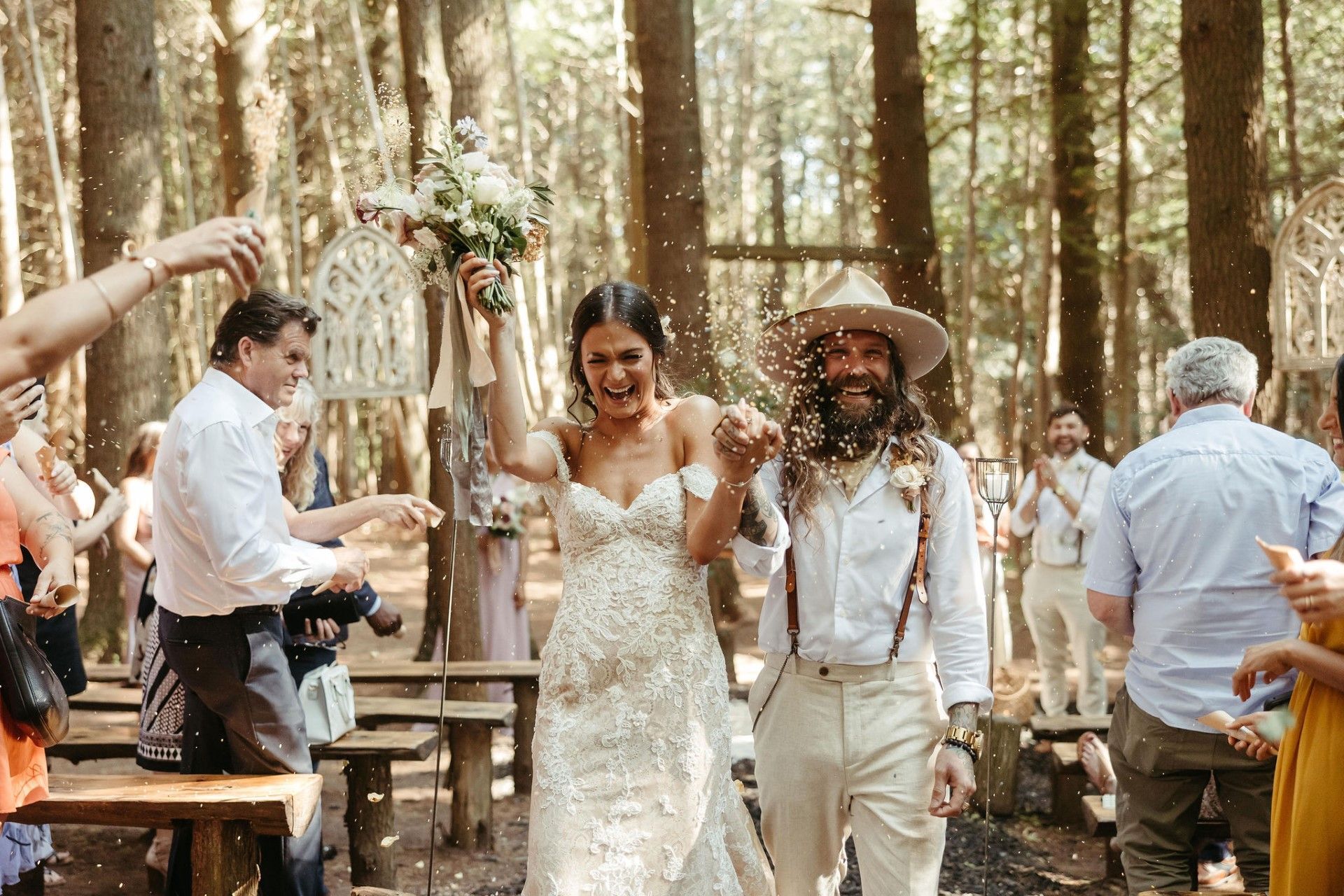 Newlyweds walk down aisle after ceremony, showered with confetti in forest setting.