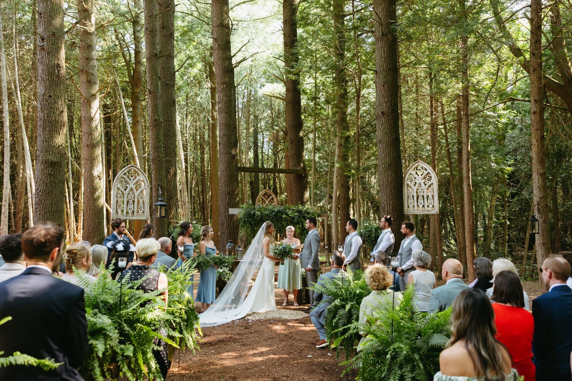 Wedding ceremony in a forest setting: Bride & groom at the altar, surrounded by guests, trees, and floral arrangements.