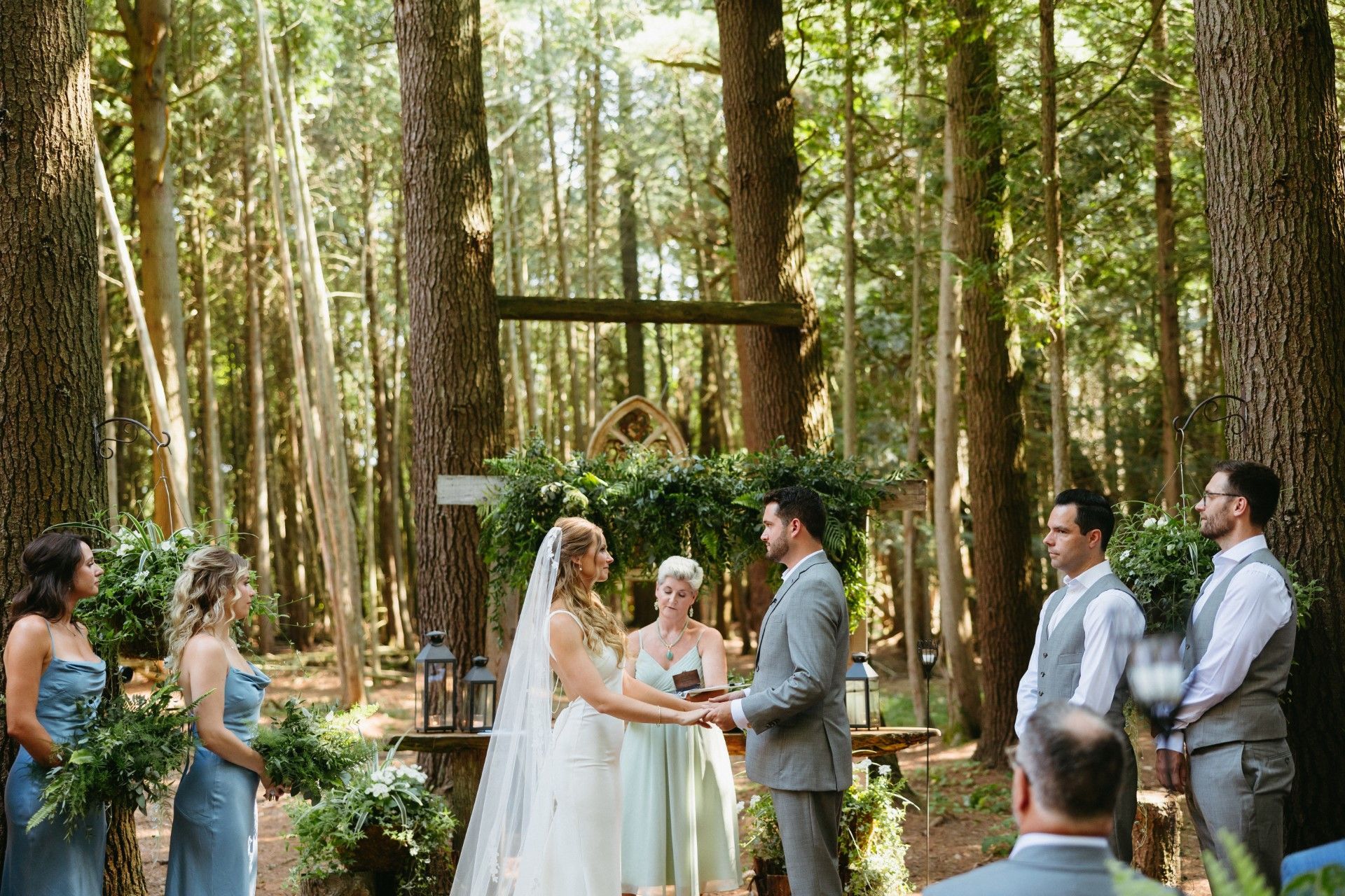 Wedding ceremony in a forest clearing. Bride and groom hold hands before an arch. Wedding party members stand by them.