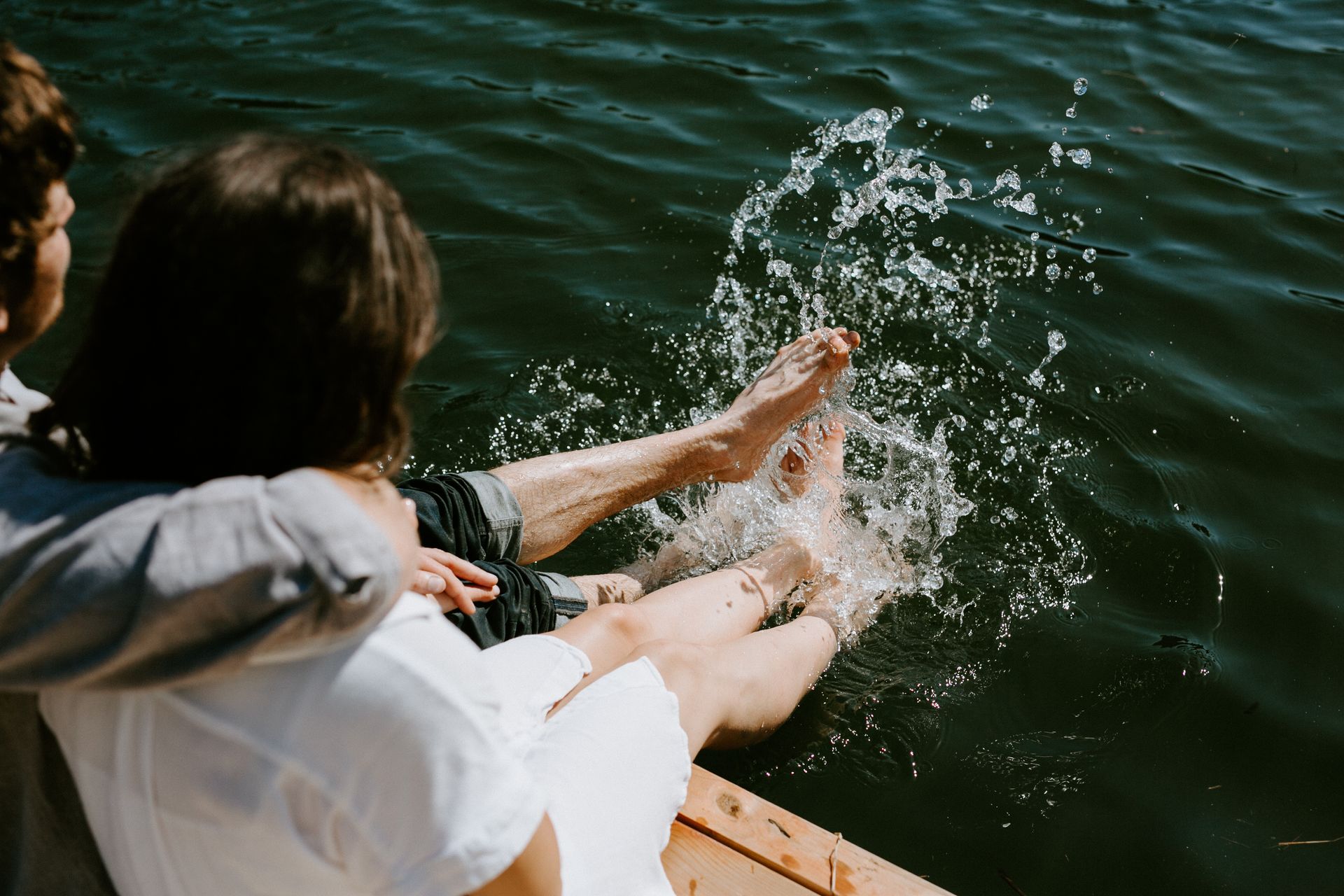 Person splashing feet in water, being embraced by another. Water sprays up, sunlight.
