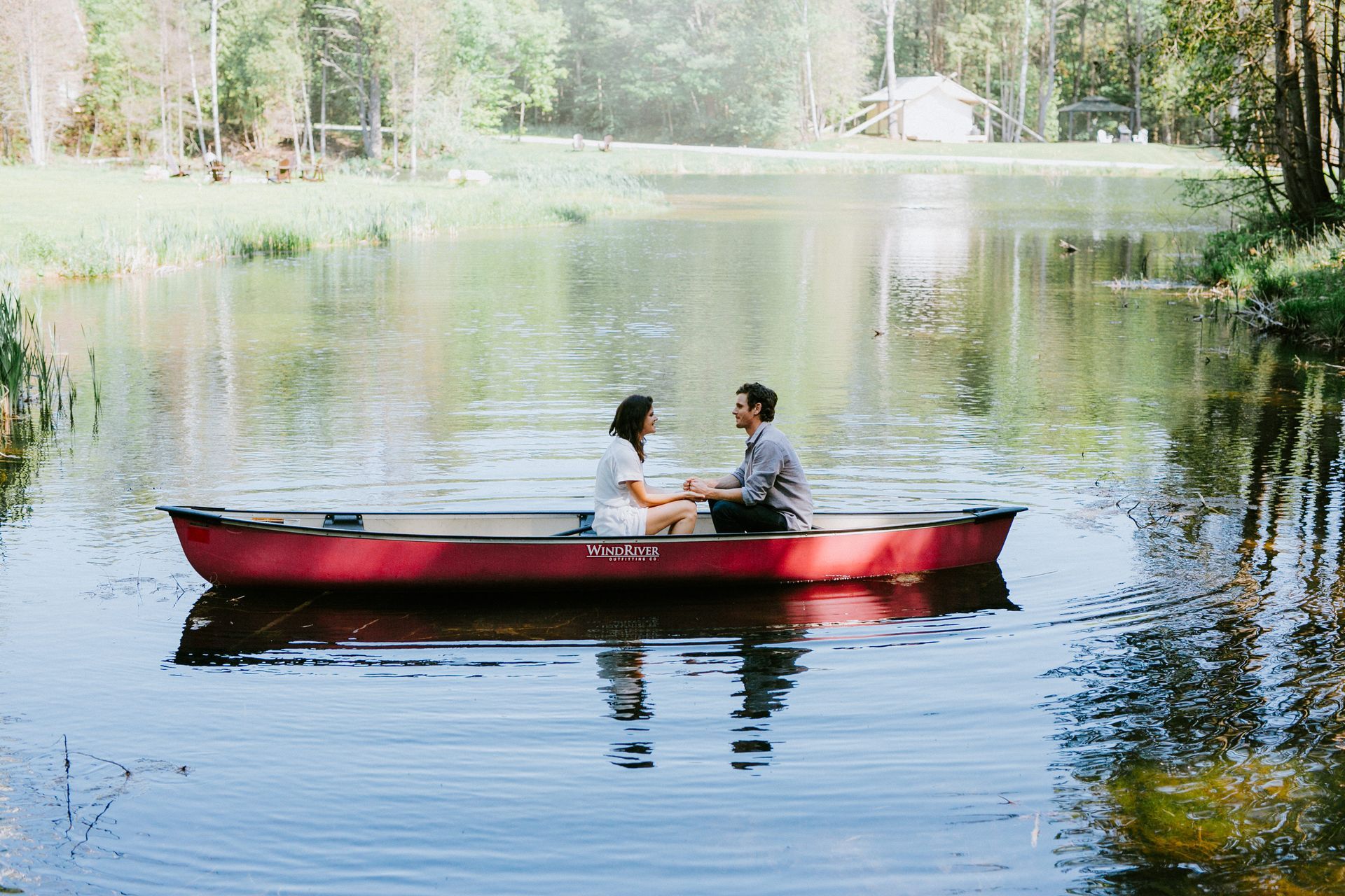 Couple in red canoe on a calm lake, holding hands, with trees in the background.