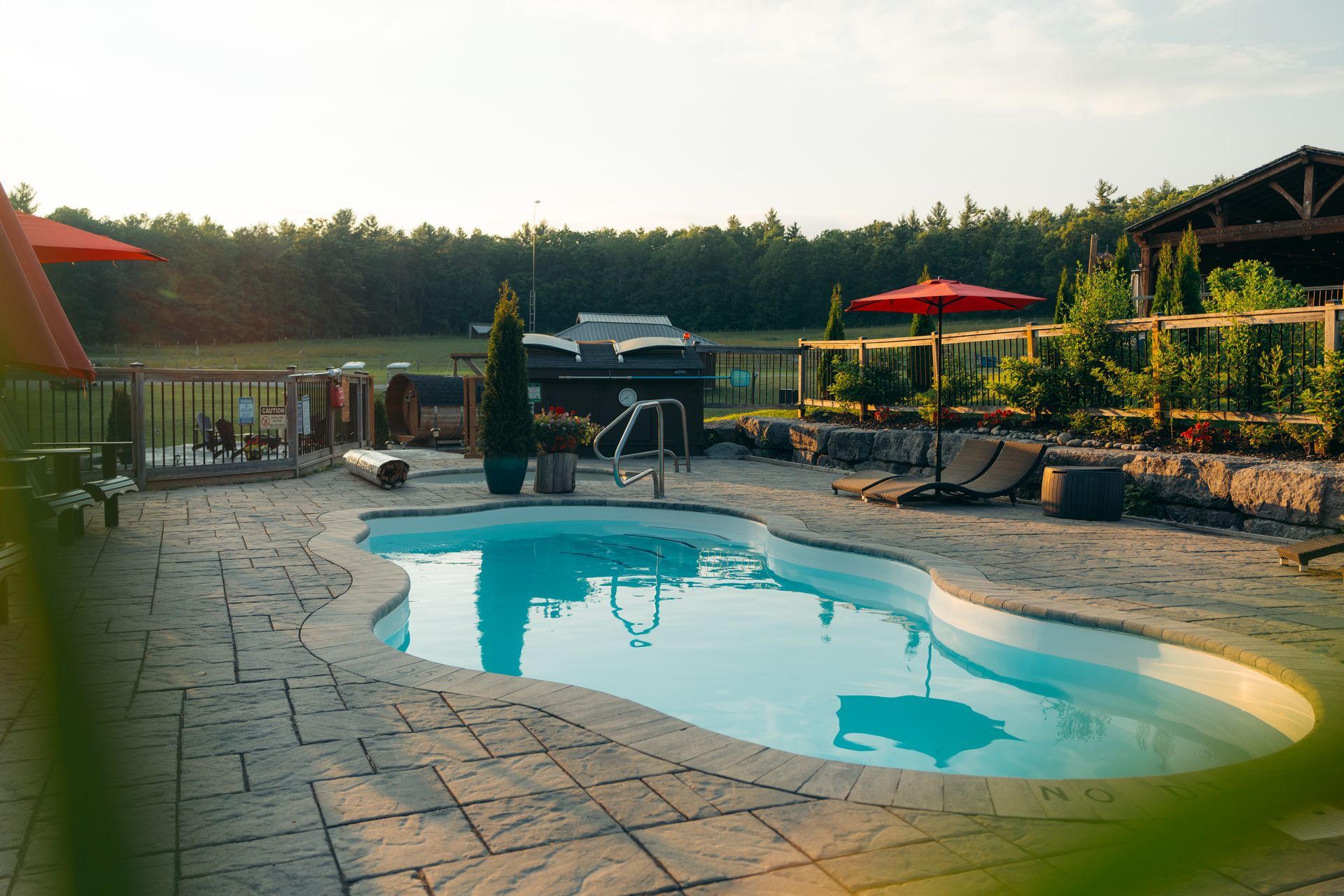 Pool with blue water surrounded by stone patio, red umbrellas, and lush greenery in a sunlit outdoor setting.