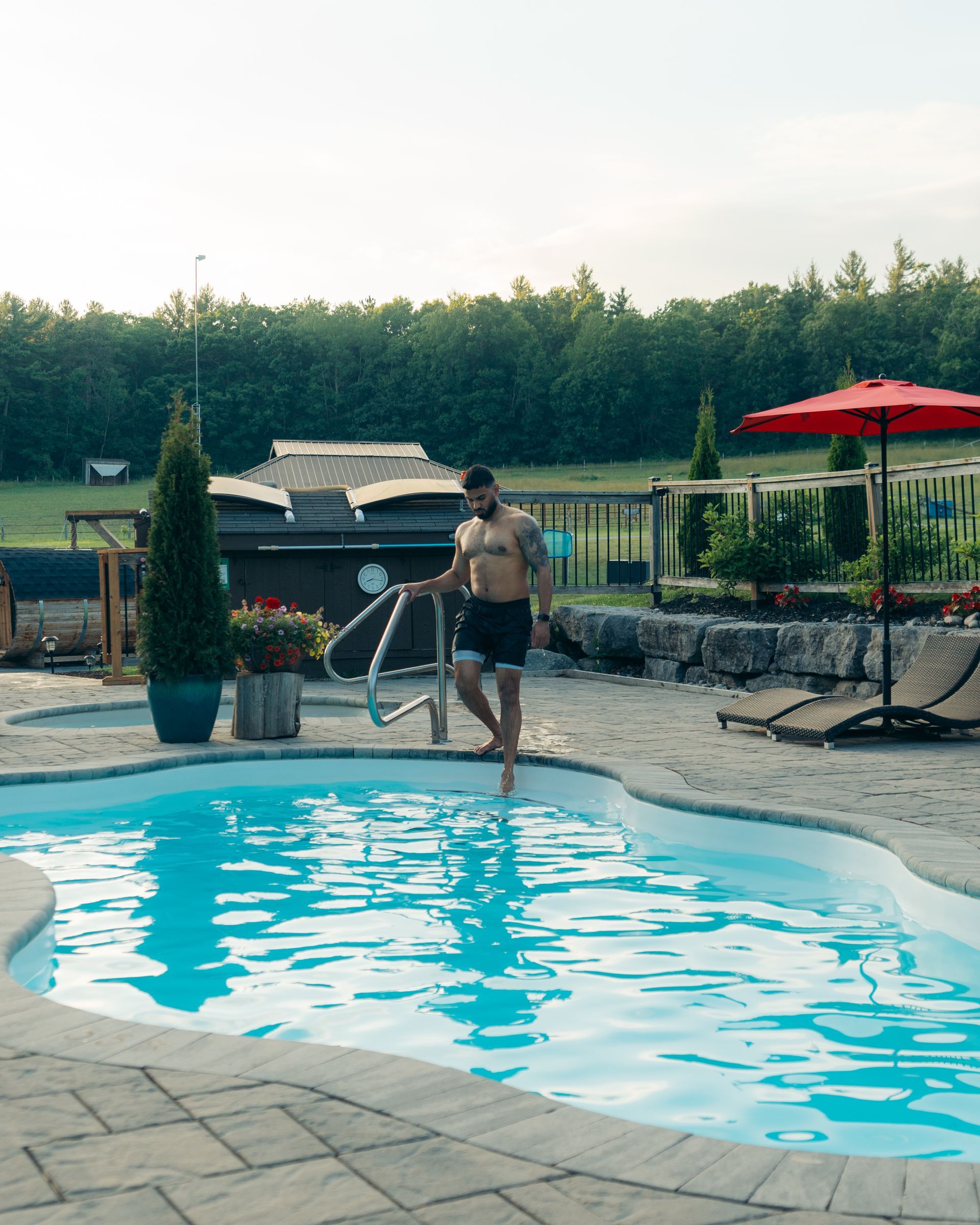 Man about to enter pool, holding handrail. Poolside setting with sun umbrella and hot tub in the background.