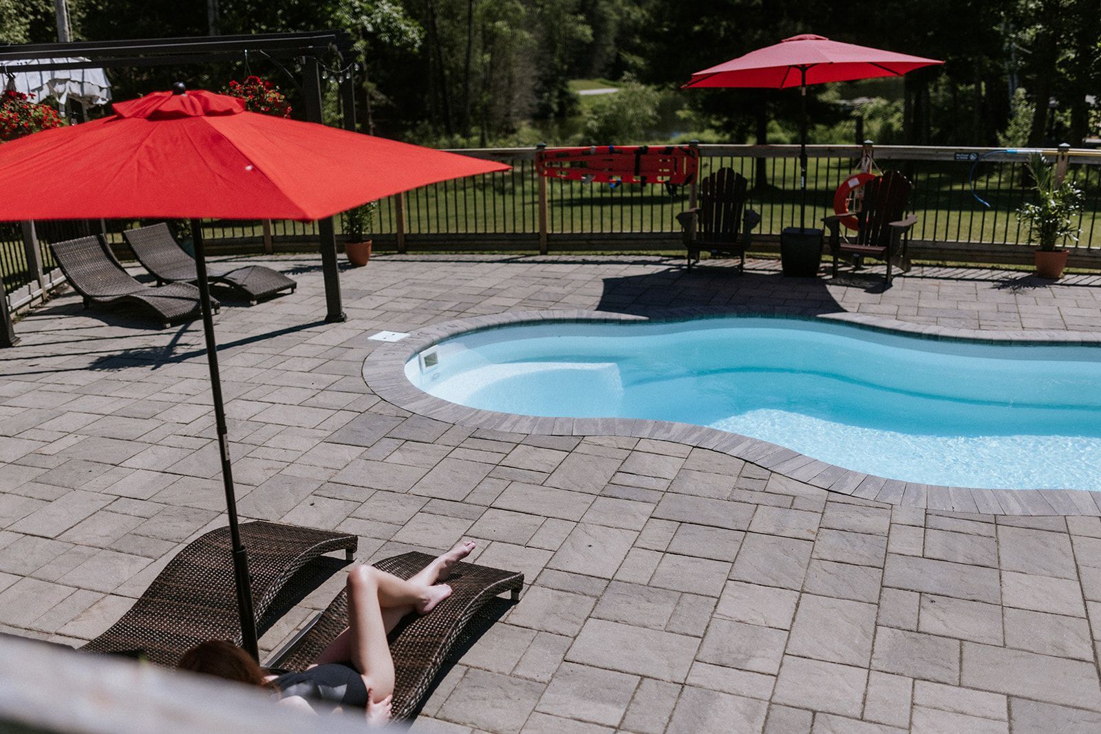 A person sunbathing by a pool with red umbrellas and lounge chairs.