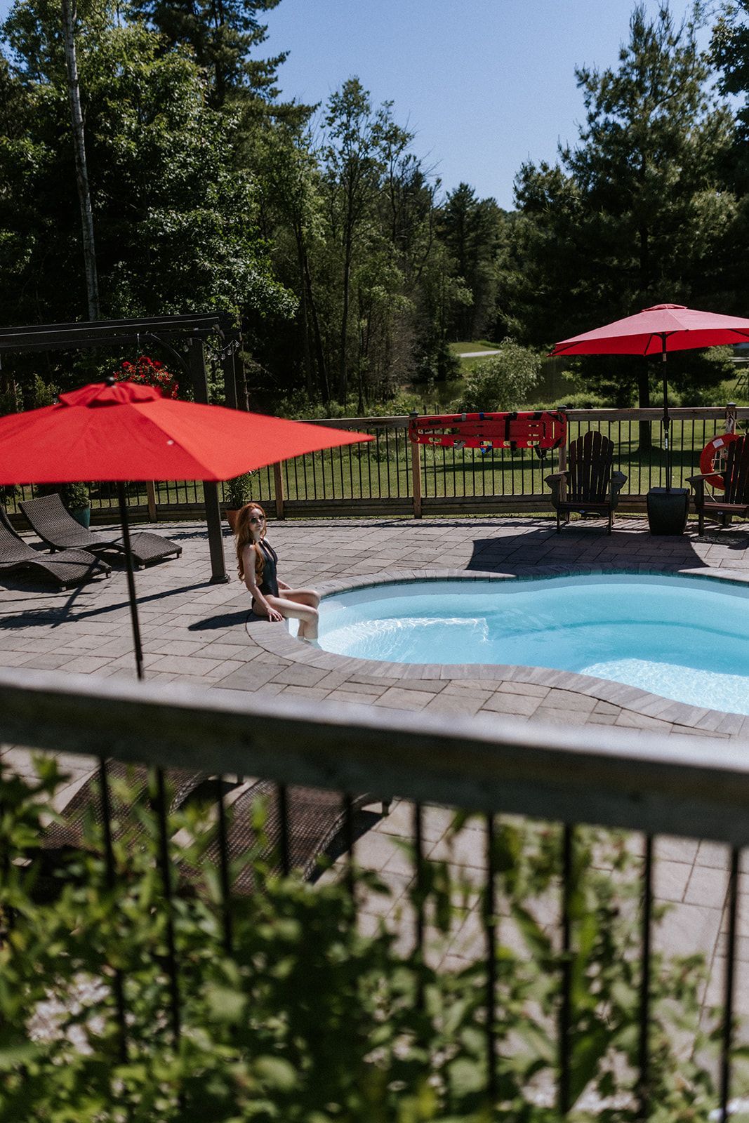 Person sitting poolside under red umbrella; pool, patio, and trees in the background.